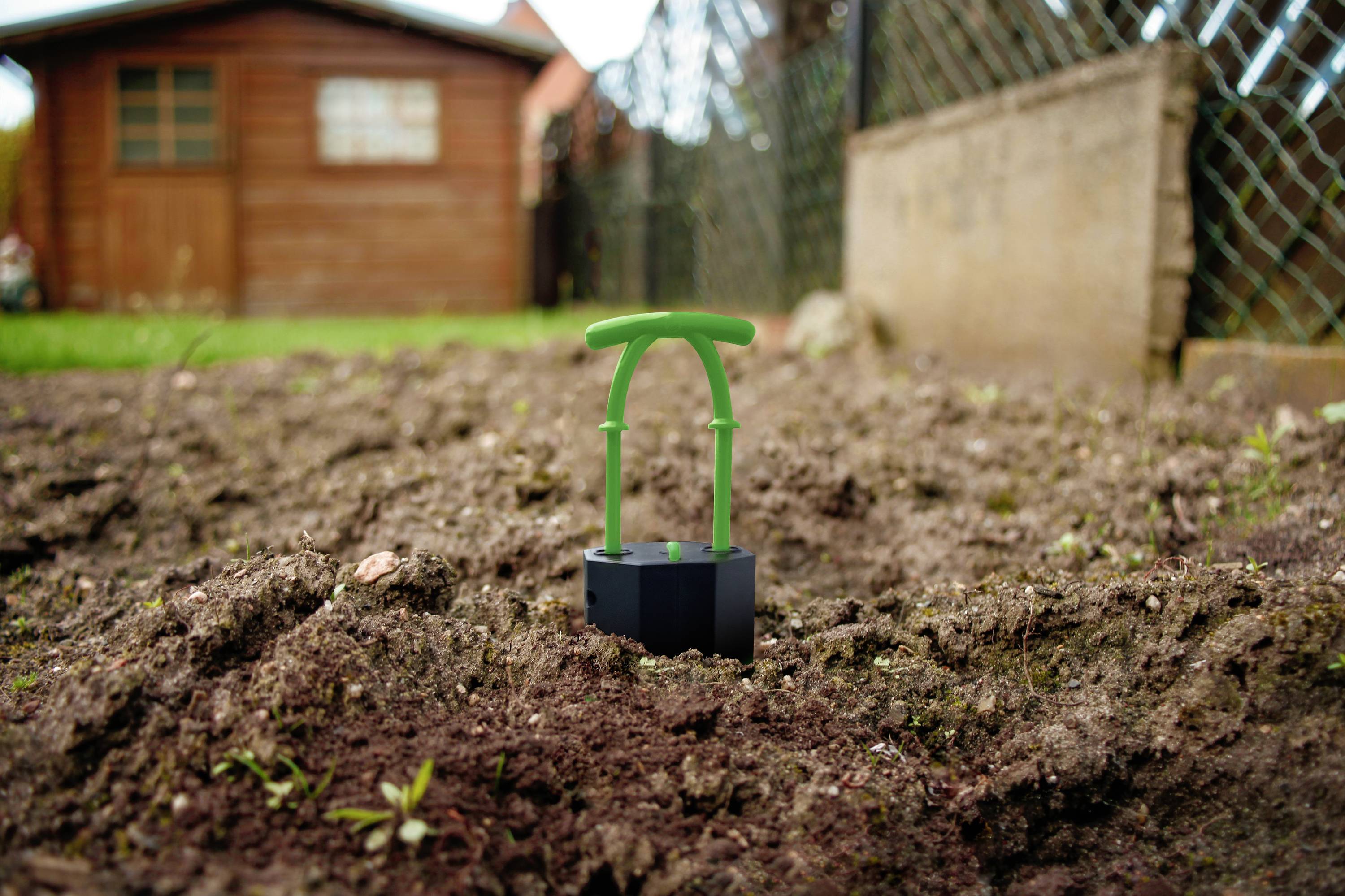Egy kerti talaj egy kis zöld ültetési segédeszközzel, amelyet a földbe szúrtak. A háttérben egy faszerszámtároló épület látható.