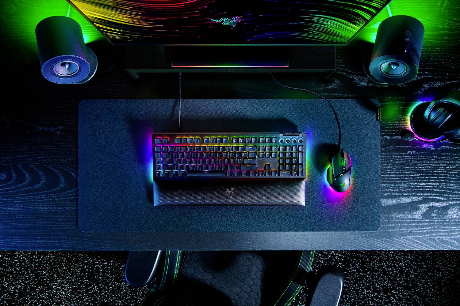 Megvilágított gaming billentyűzet és egér egy fekete íróasztalon, hangszórókkal és monitorral, színes LED-fények körülvéve.