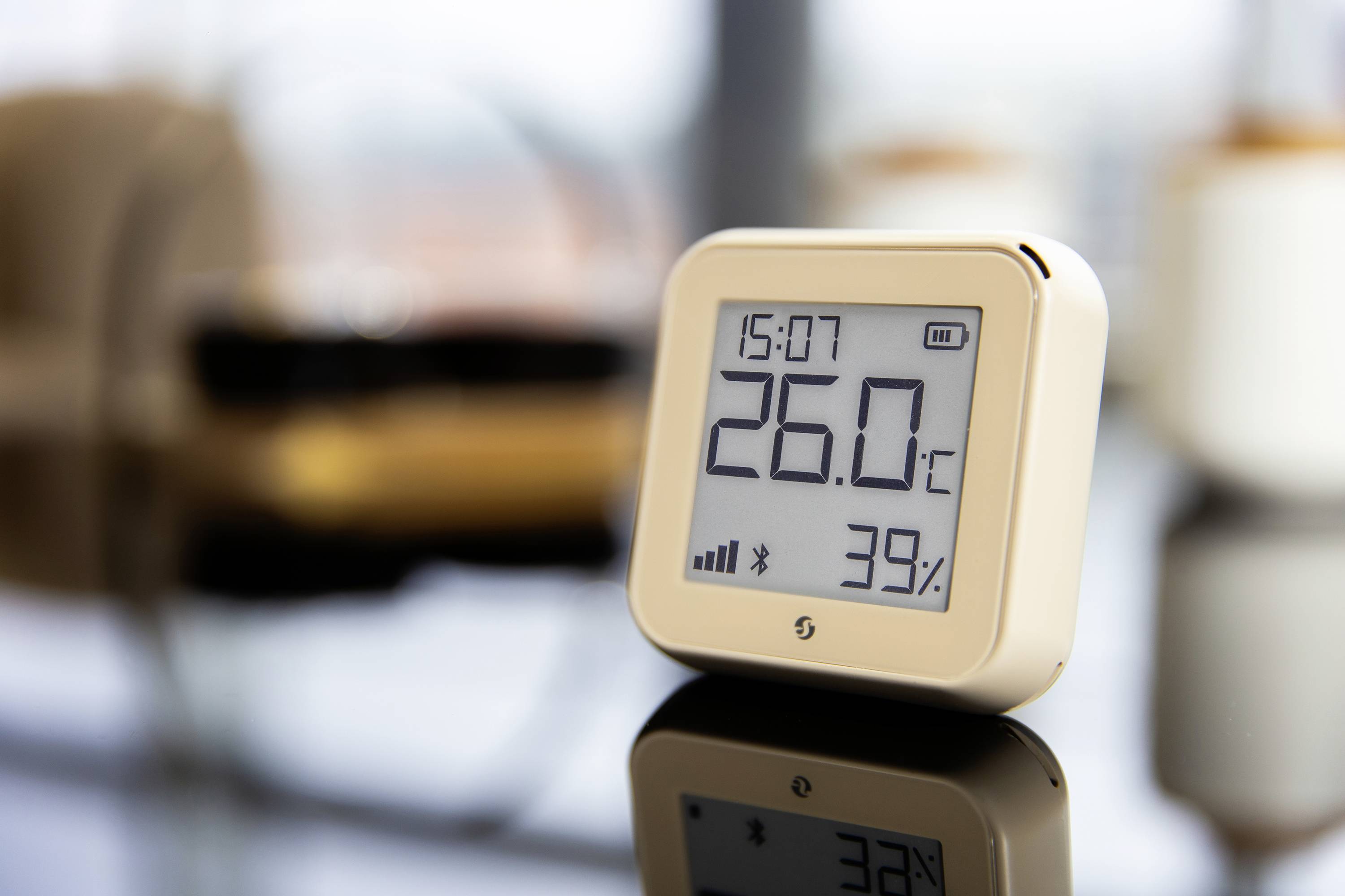 Egy digitális hőmérő 26,0°C-ot és 39% páratartalmat mutat. Egy tükröződő felületen áll egy modern helyiségben.