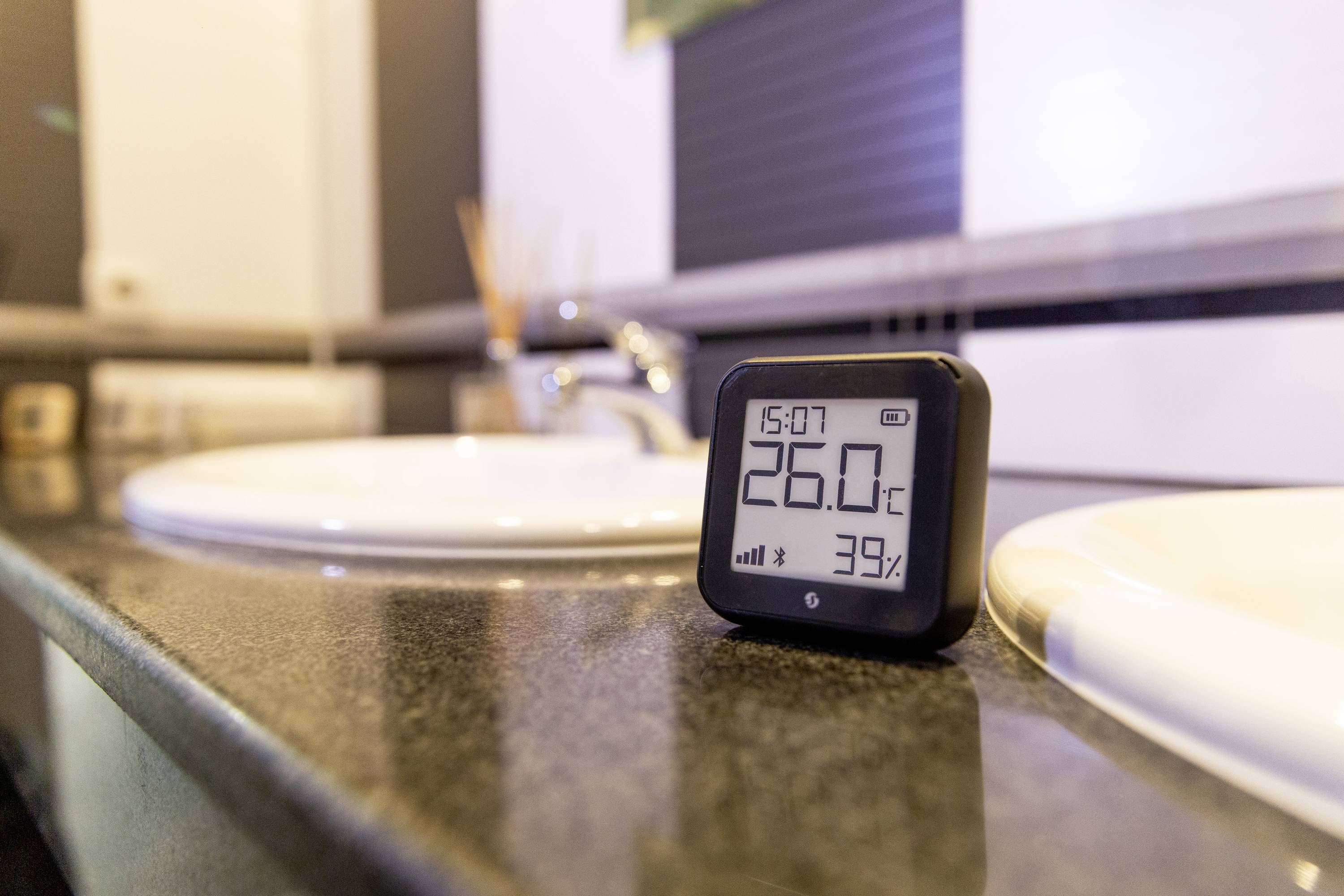 Egy digitális hőmérő 26,0°C-ot és 39% páratartalmat mutat egy fürdőszobai mosdópulton.