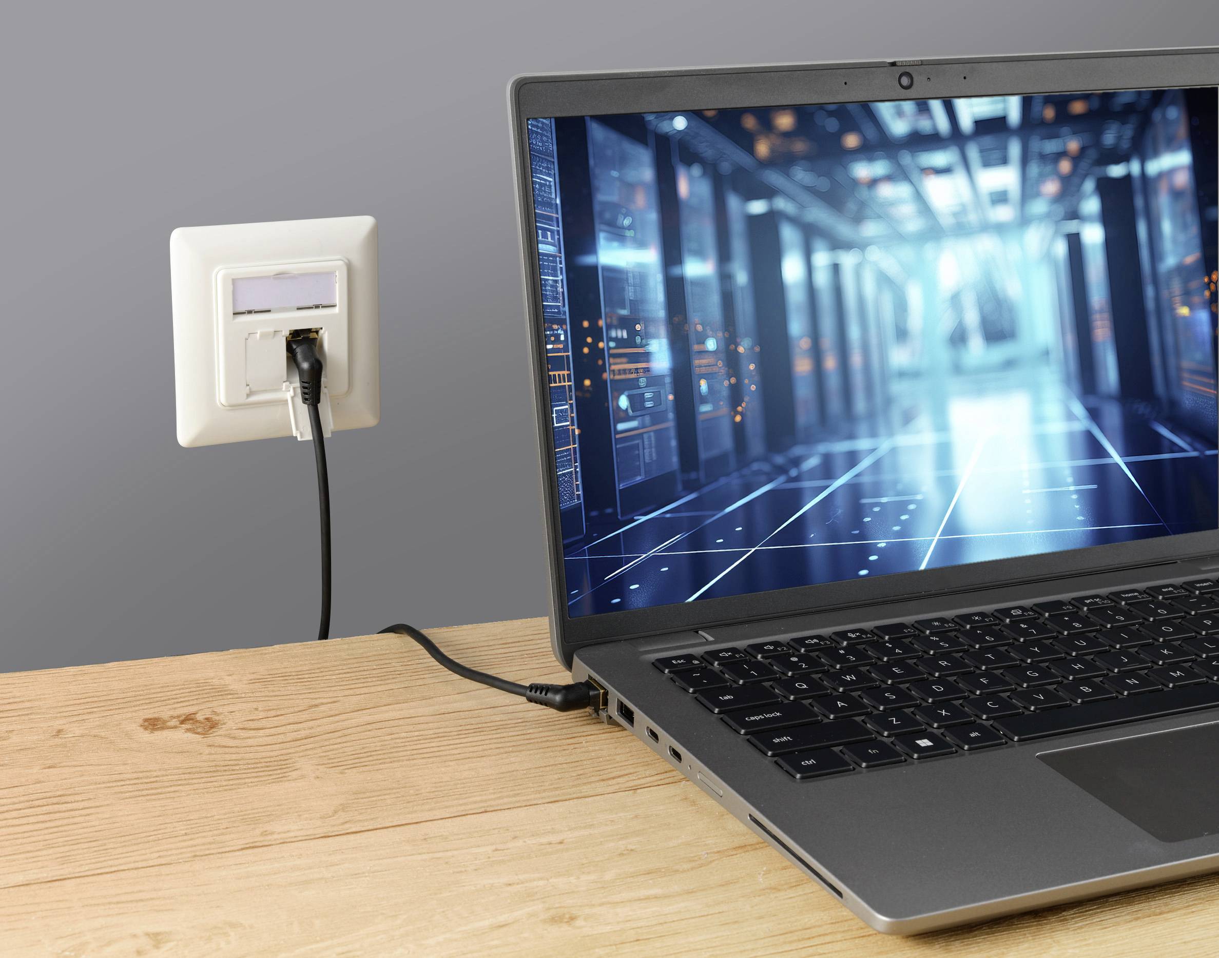 Egy laptop fából készült asztalon egy digitális adatközpontot mutat a képernyőjén, amelyet a falban lévő hálózati aljzattal összekapcsolva látunk.