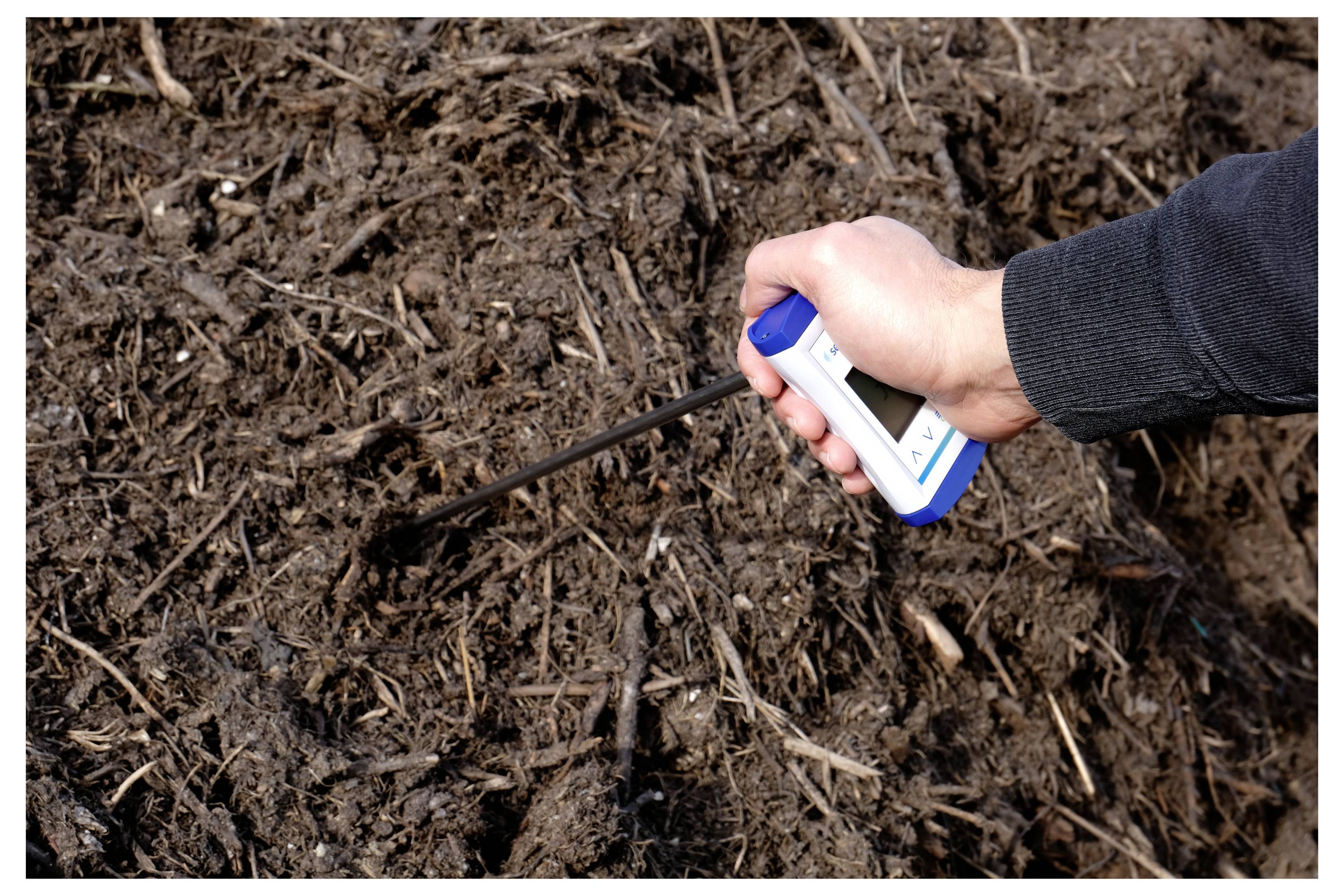 Egy személy keze digitális hőmérővel méri a sötét, lebomló talaj vagy komposzt hőmérsékletét.