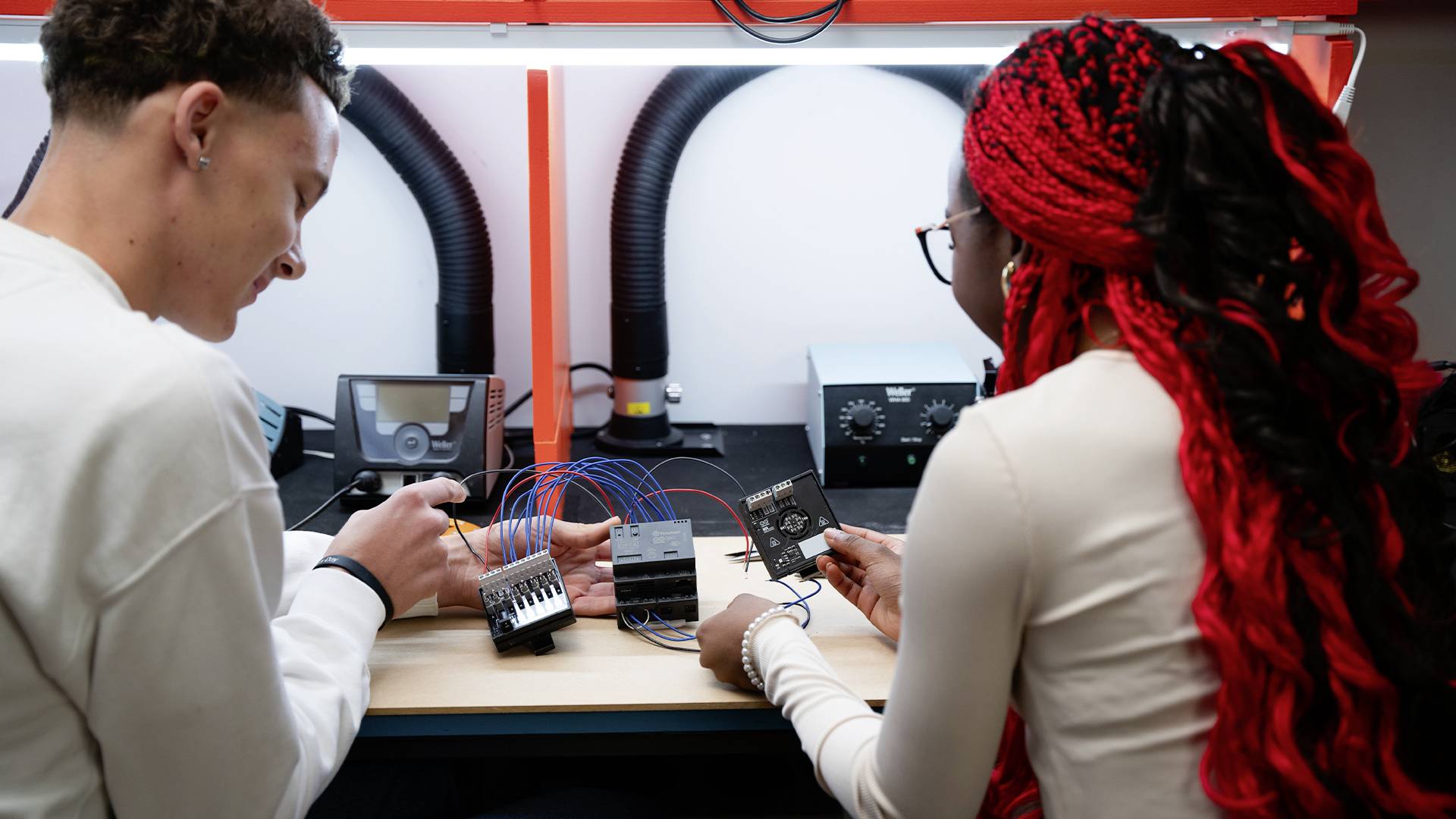 Két személy együtt dolgozik egy elektronikai laborban, egy projekten, kábelek és áramkörök között egy asztalon.