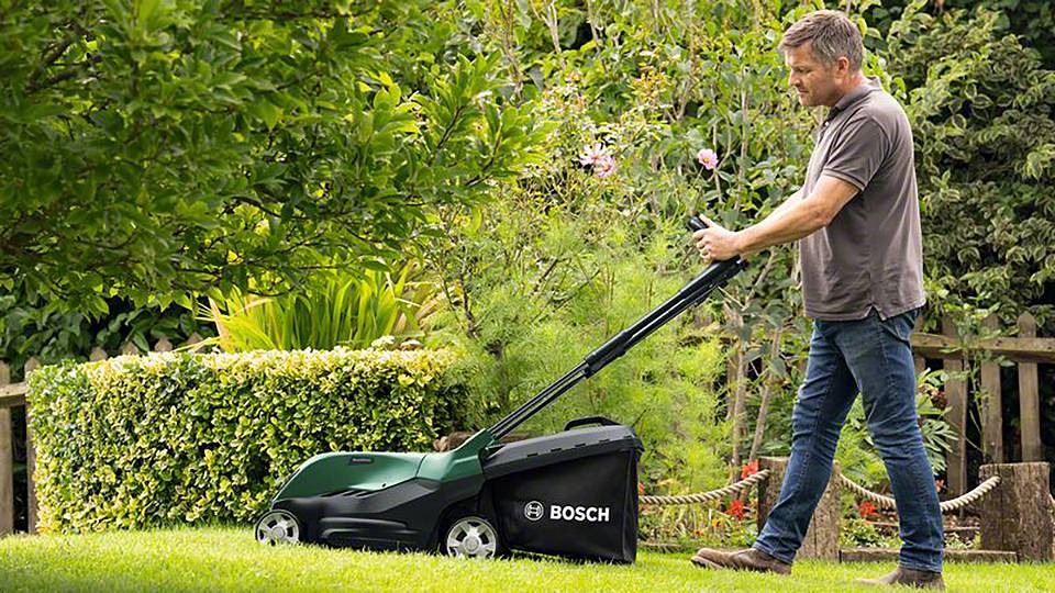 Egy férfi Bosch fűnyíróval nyírja a füvet egy zöld kertben, növények és egy fa kerítés övezik a háttérben.
