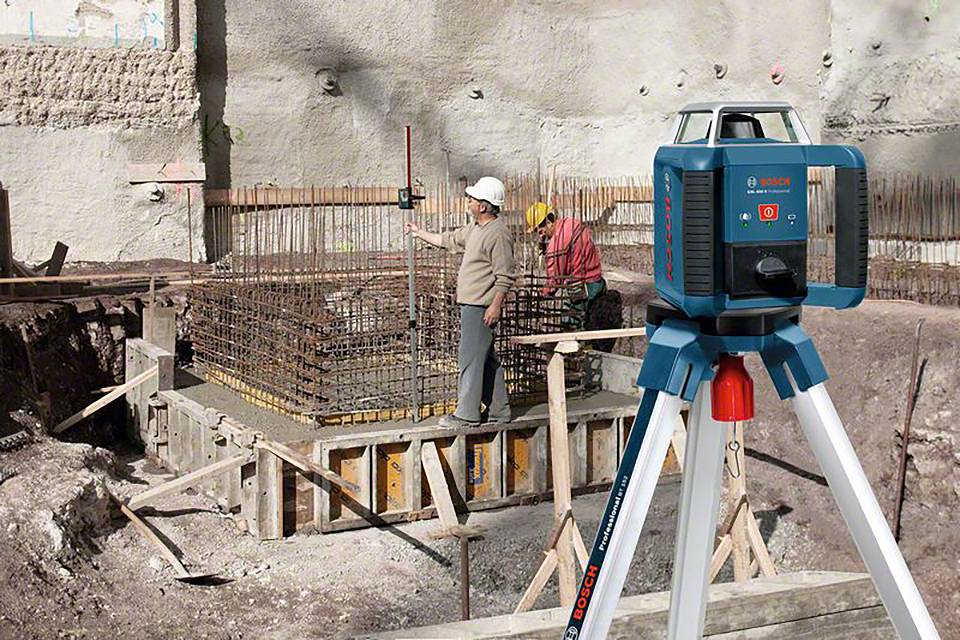 Két munkás dolgozik egy építkezésen, előtérben egy lézeres mérőműszerrel. A betonzsaluzatot mérik fel.