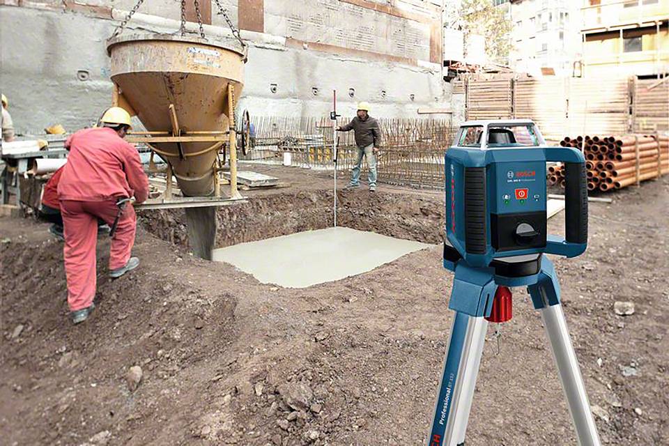 Építőmunkások betonnal töltenek fel egy alapgödröt egy építkezésen, miközben egy lézeres szintező műszer állványon van beállítva.