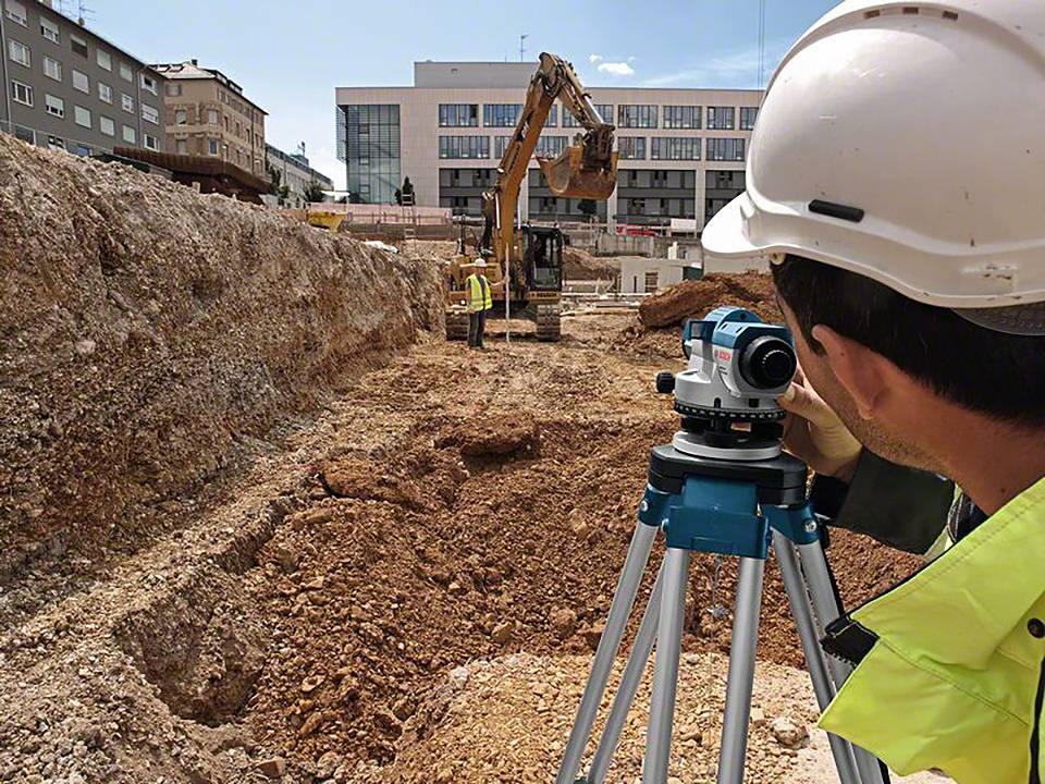 Munkások egy építkezésen, előtérben egy földmérő mérnök teodolittal, háttérben egy kotró, épületek övezik a helyszínt.