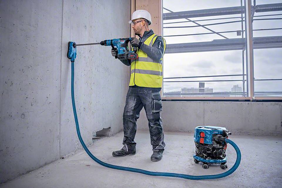 Egy munkás védősisakban és biztonsági mellényben furatot készít egy betonfalba egy építkezésen. Mellette egy porszívó áll.