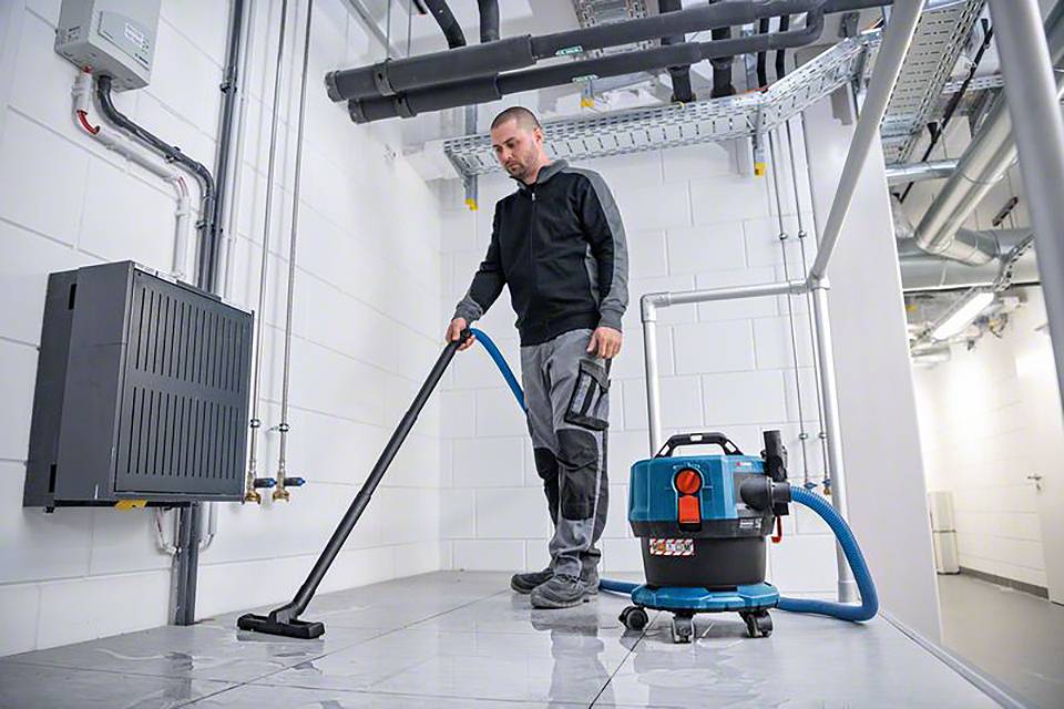 Egy férfi egy ipari porszívóval takarítja a padlót egy technikai helyiségben. A falon csövek és egy elosztószekrény látható.