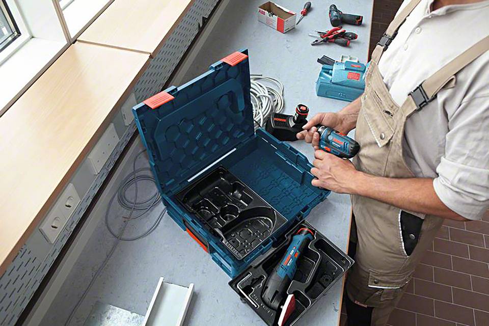 Egy szerelőruhát viselő személy elektromos szerszámot tart a kezében, egy nyitott szerszámosláda előtt állva, amelyben több eszköz is látható.