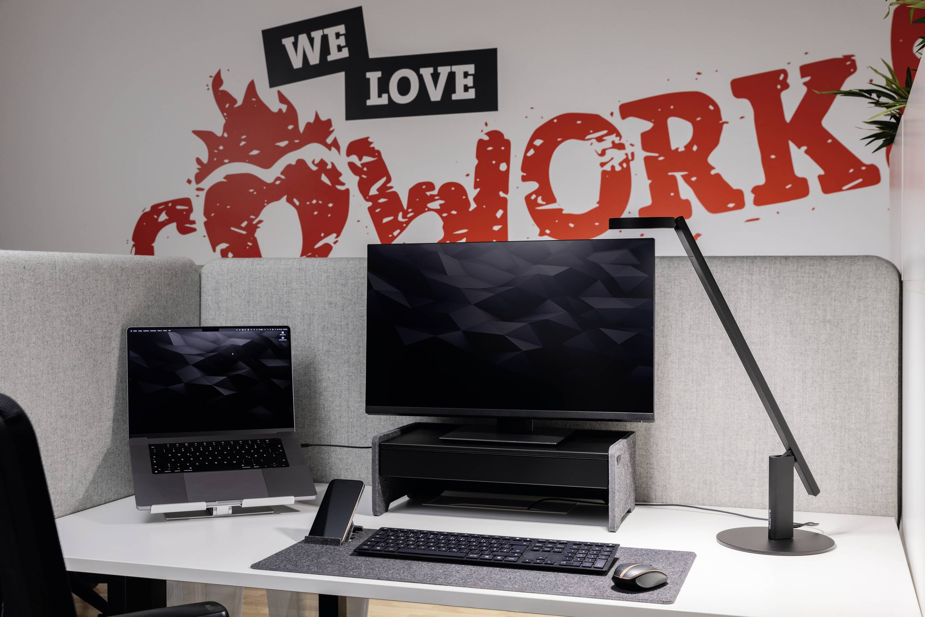 Egy modern munkahely laptop, monitor, lámpa és a háttérben a falon elhelyezett 'We love cowork' felirattal.
