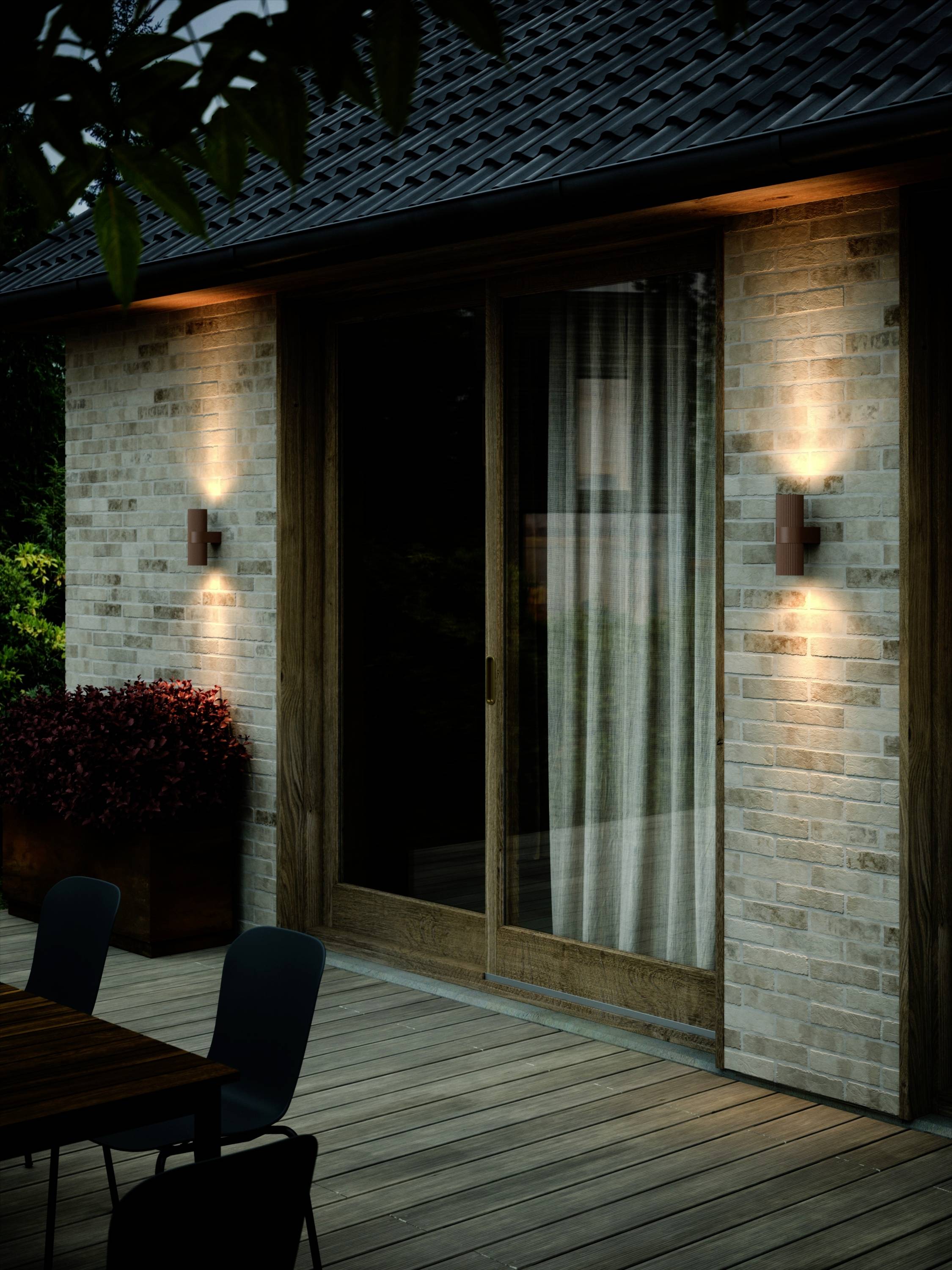 Egy megvilágított külső tér, amelyen terasz található székekkel és egy asztallal, téglafal előtt tolóajtókkal.