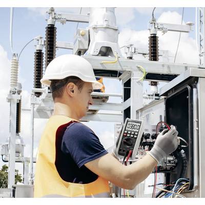 Egy technikus védősisakban és biztonsági mellényben multiméterrel ellenőriz elektromos berendezéseket egy szabadtéri villamos alállomáson.