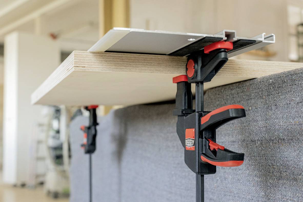 Egy szorító egy fadarabot és egy fehér lapot tart együtt egy munkapadon. A jelenet egy műhelyben játszódik.