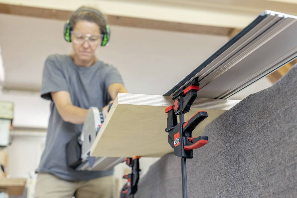 Egy személy egy fűrésszel fadeszkát vág. A deszkát szorítóbilincsekkel rögzítik. A személy védőszemüveget és fülvédőt visel.