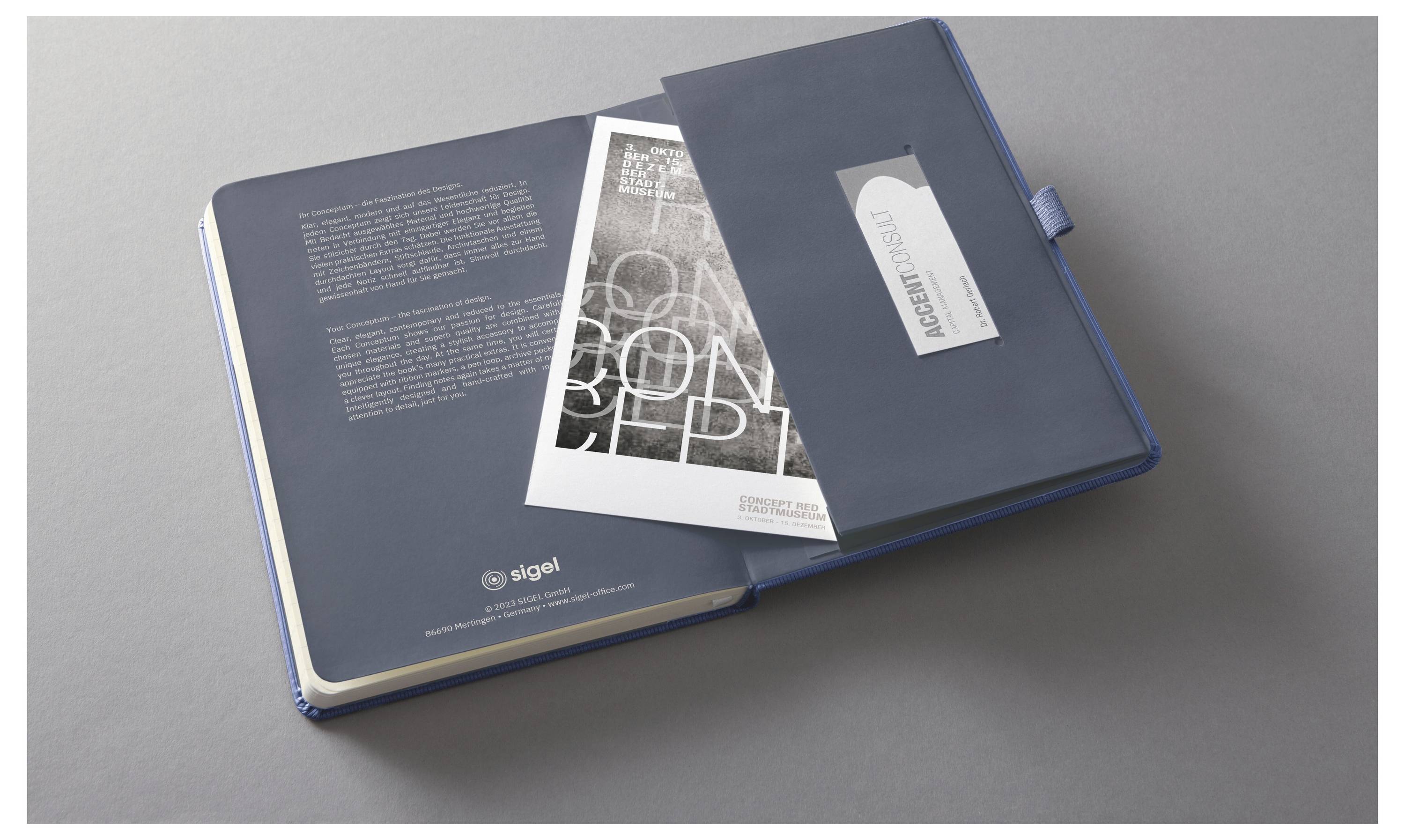 Nyitott kék jegyzetfüzet vagy tervező egy szürke felületen, amely nyomtatott lapokat, egy borító fülét és egy "PROJECT BOOK" feliratú zseb lapot mutat.