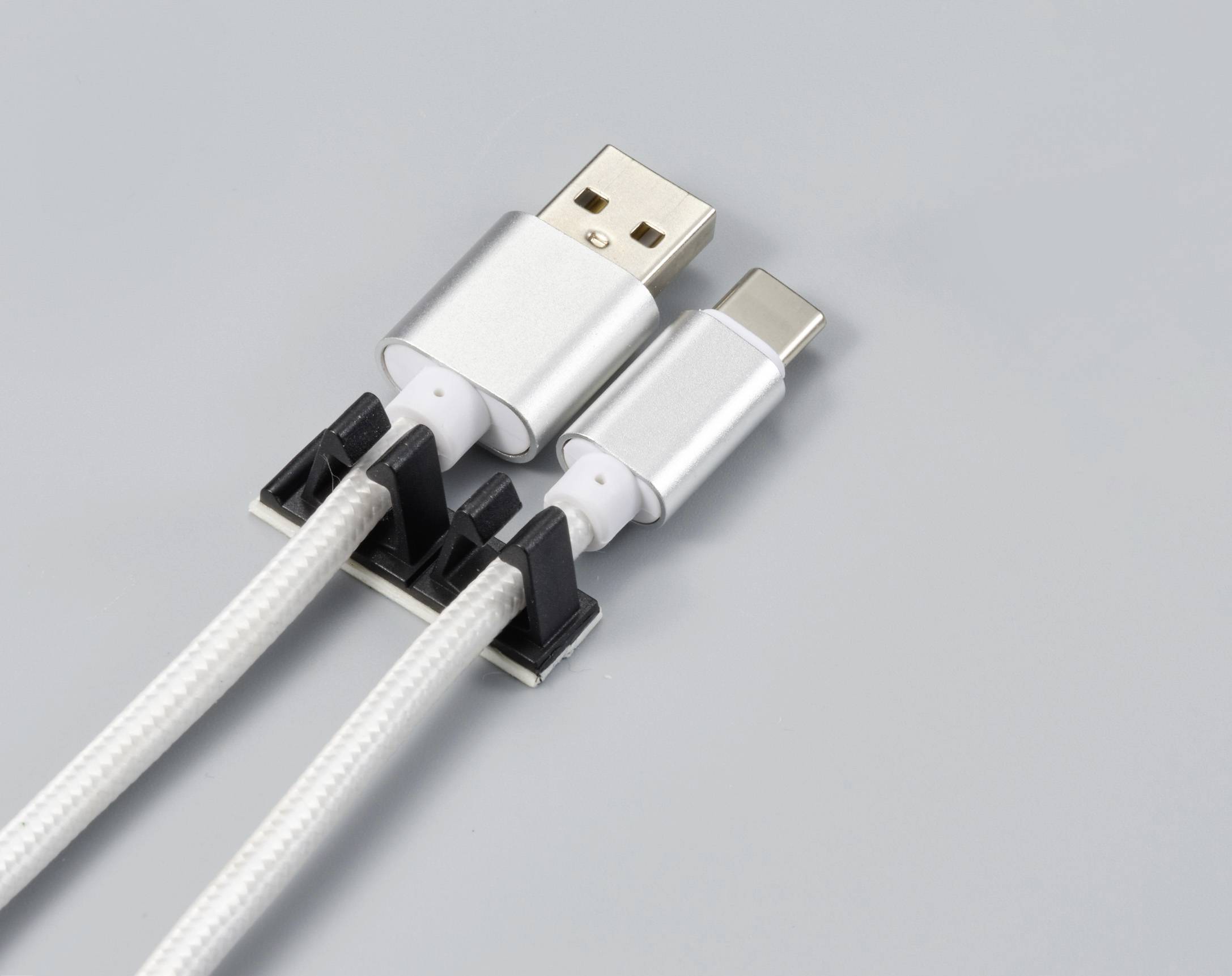 Egy fekete kábelrendező által egy szürke háttéren rögzített két egymás melletti ezüstszínű kábel, USB- és USB-C-csatlakozókkal.