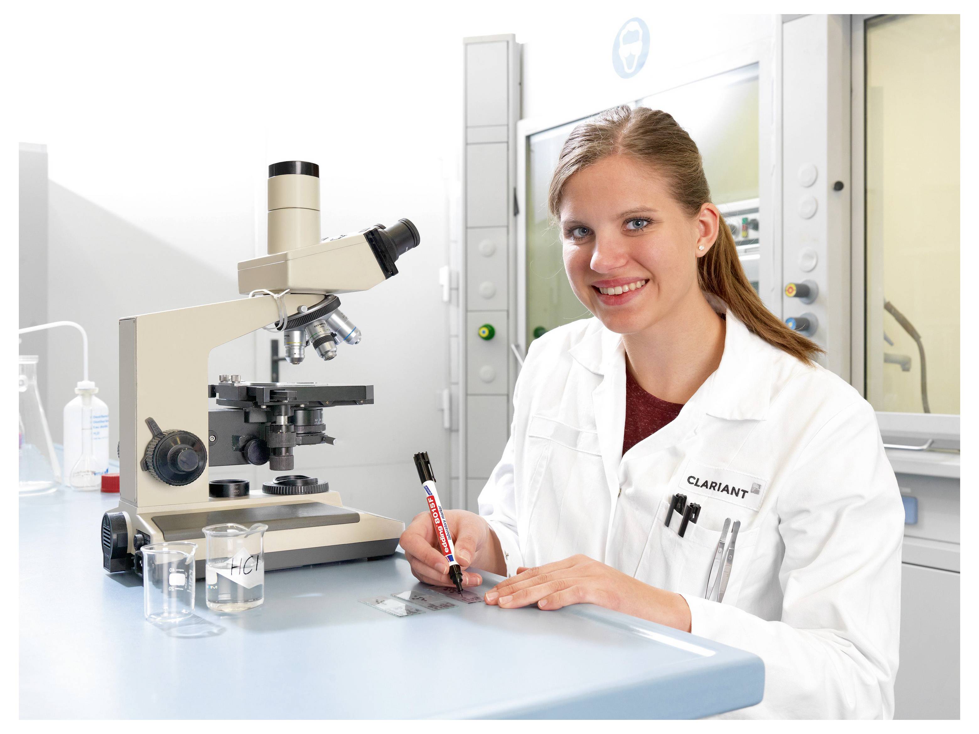 Mosolygó női tudós fehér laboratóriumi köpenyben ír egy laboratóriumi asztalnál, amelynek mellett mikroszkóp és üvegpoharak vannak egy fényes laboratóriumban.