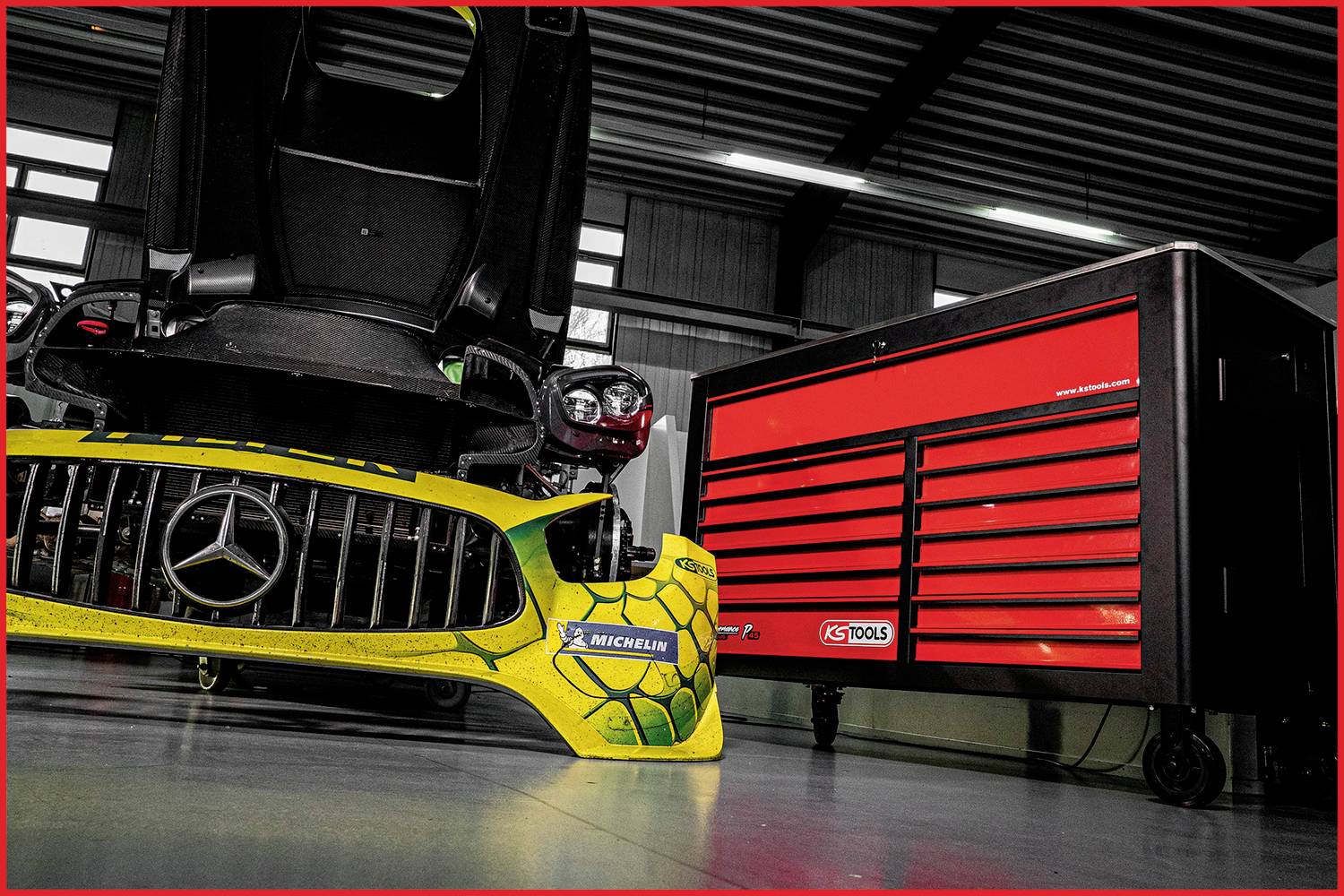Egy sárga Mercedes autó lökhárítója fekszik egy piros szerszámszekrény mellett egy műhelyben. Felül lámpák és ablakok láthatók.