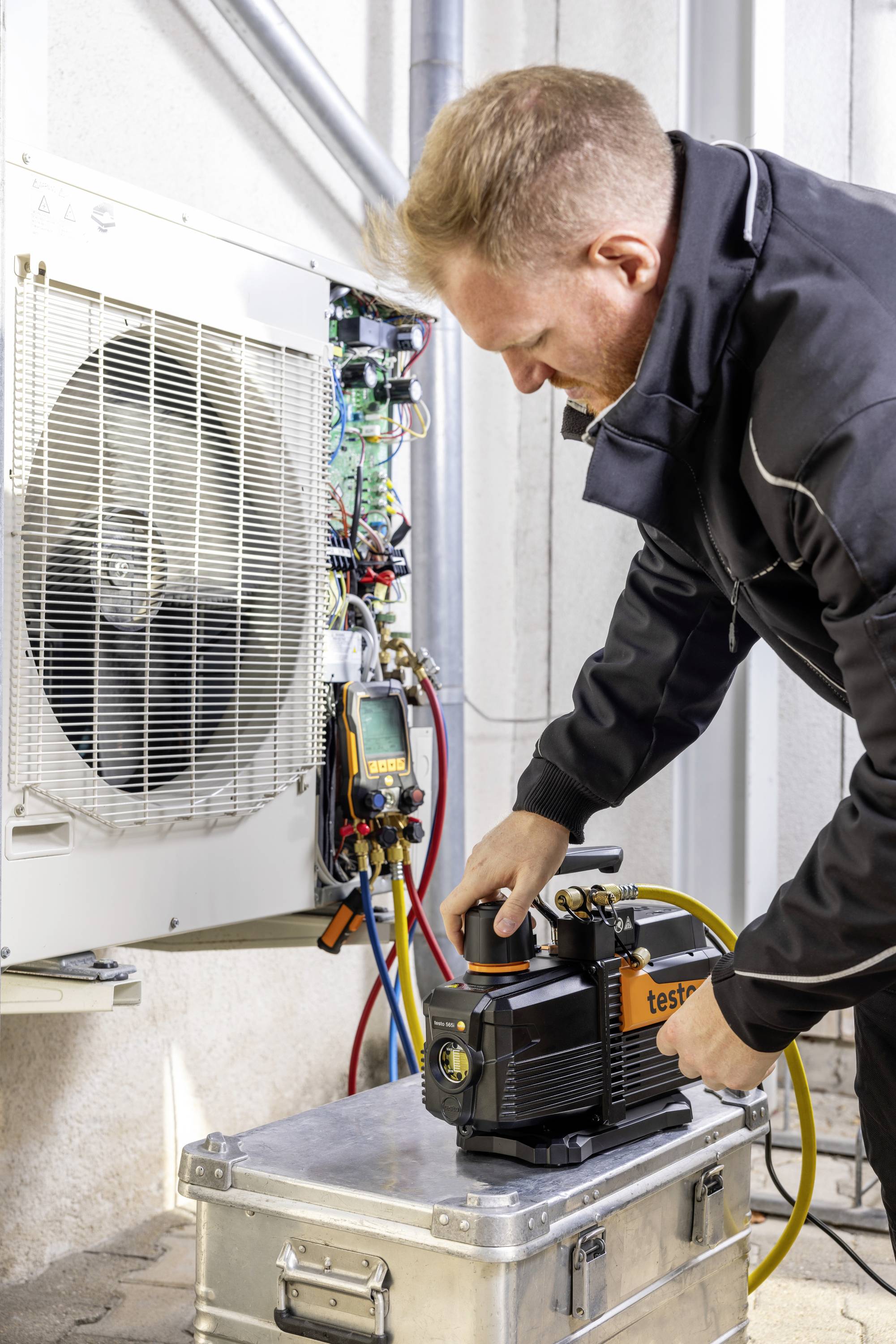 Egy technikus karbantartja a klímaberendezést, műszerekkel felügyeli annak működését és ellenőrzi a csatlakozásokat egy fémdobozon.