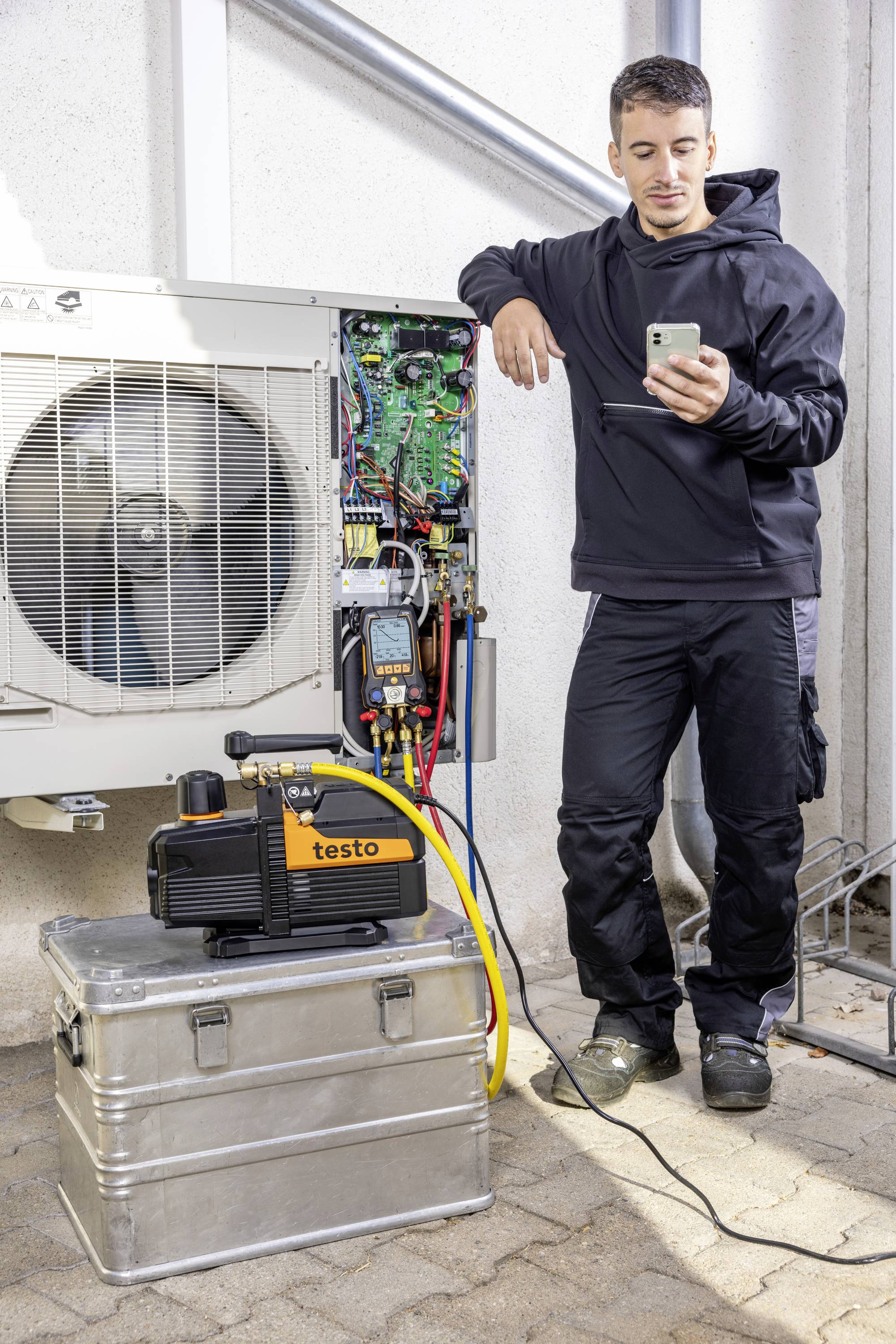 Egy férfi egy okostelefon segítségével ellenőriz egy légkondicionáló rendszert. Mellette egy 'Testo' márkájú szivattyú áll egy fémdobozon.