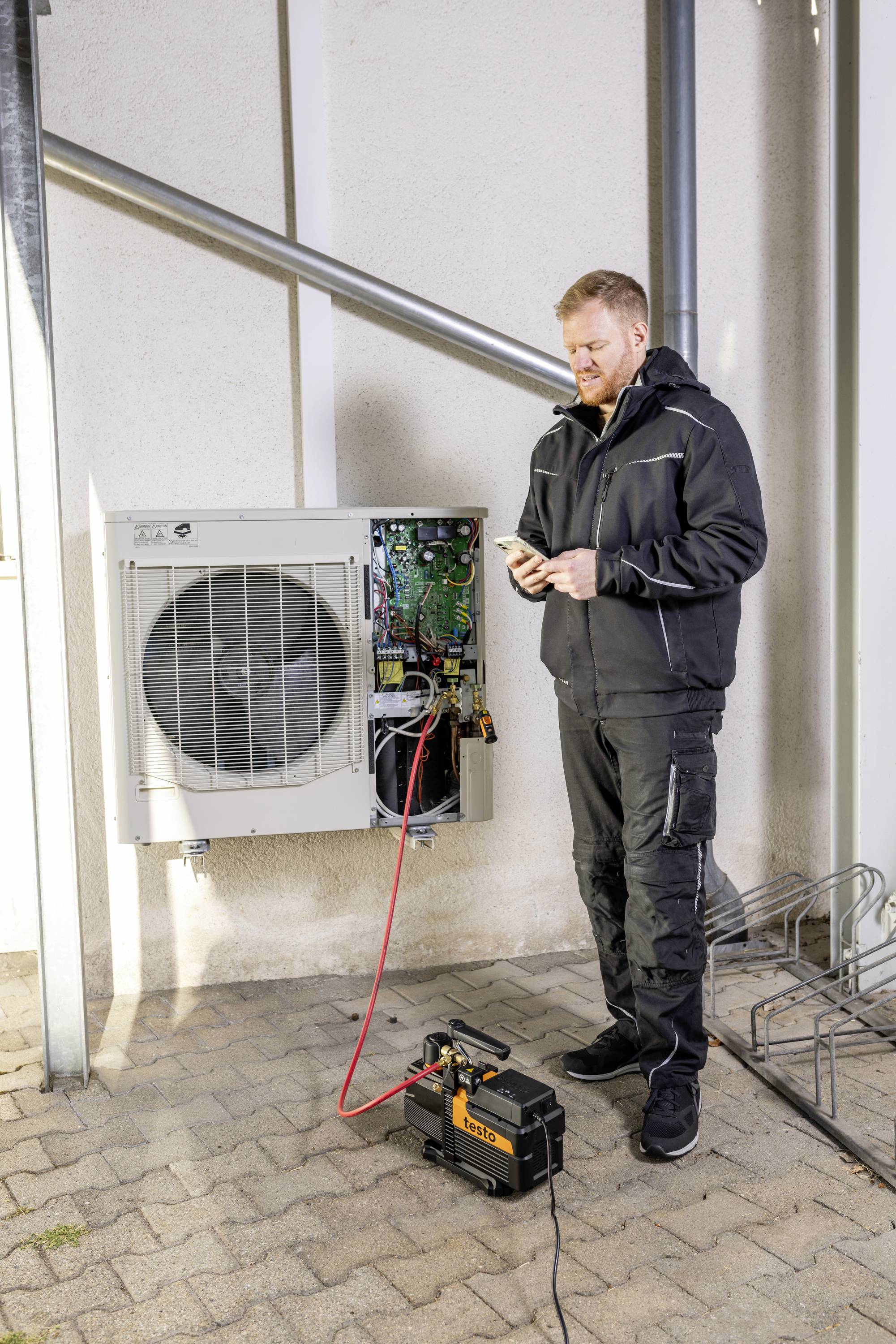 Egy technikus kint javít egy légkondicionálót. Egy szerszámot tart a kezében, és egy nyitott készülék mellett áll, amelyből kábelek lógnak ki.