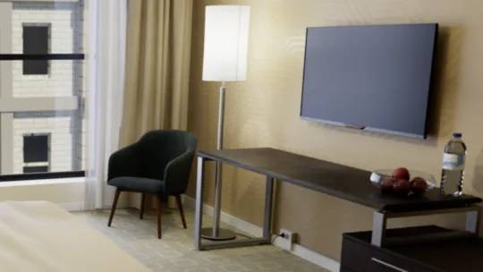 Egy modern hotelszoba egyággyal, íróasztallal, székkel és lapos képernyős televízióval. Ablak kilátással az épületekre.