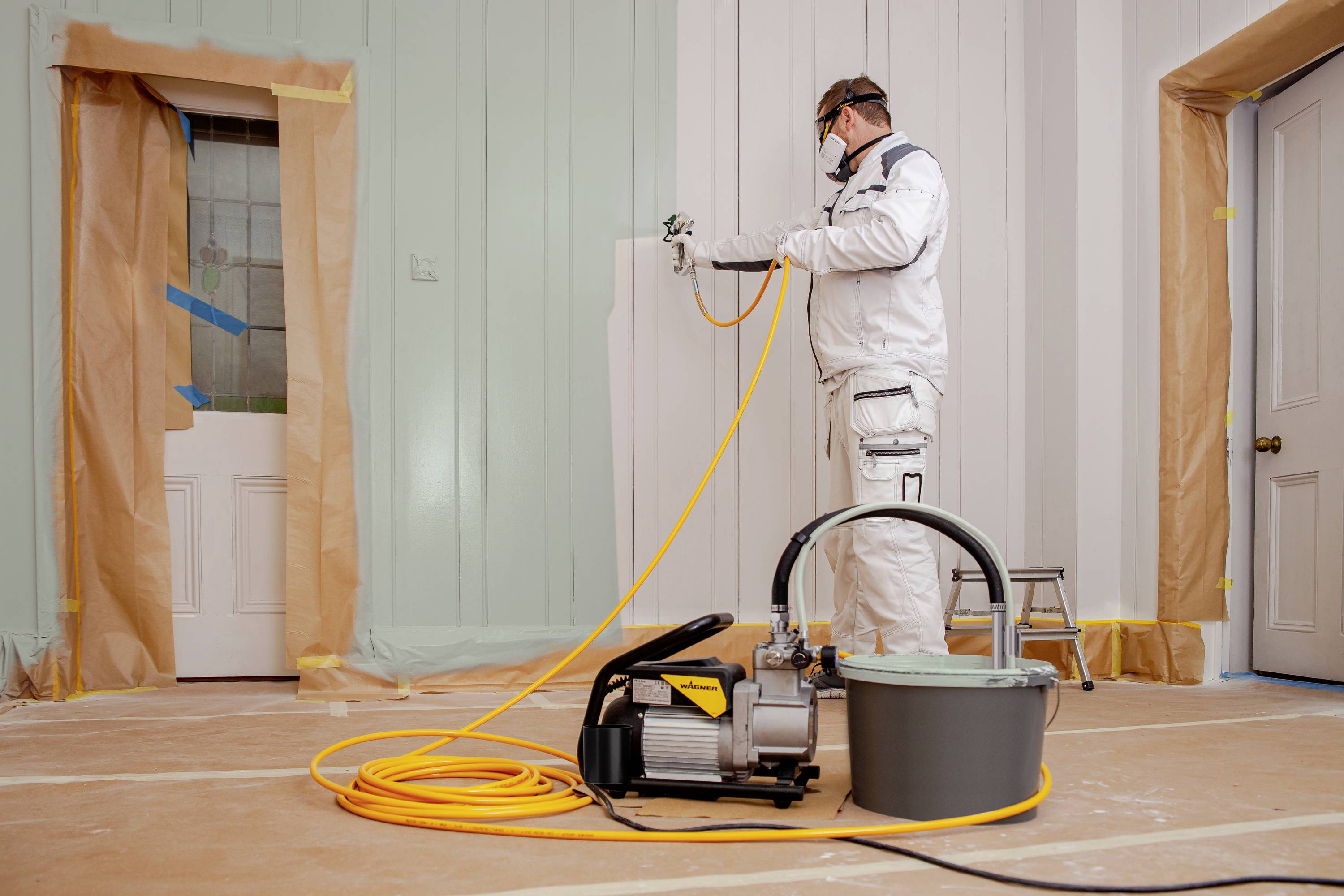 Egy festő védőruhában és szájmaszkban festékszóróval fest falakat egy fóliával letakart helyiségben.