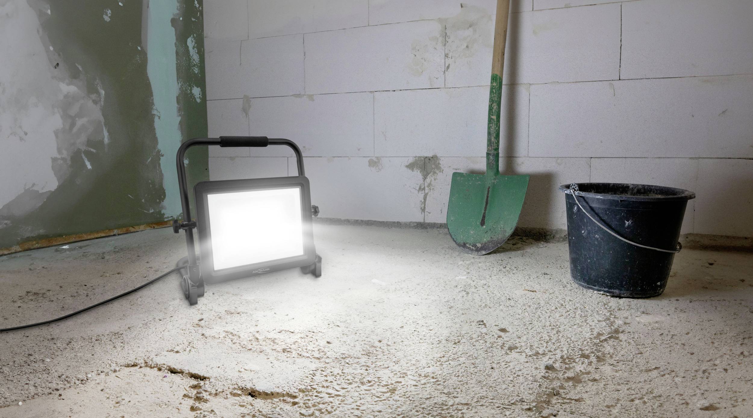 Építési munkaterület világos reflektorral, zöld ásóval és fekete vödörrel betonpadlón.