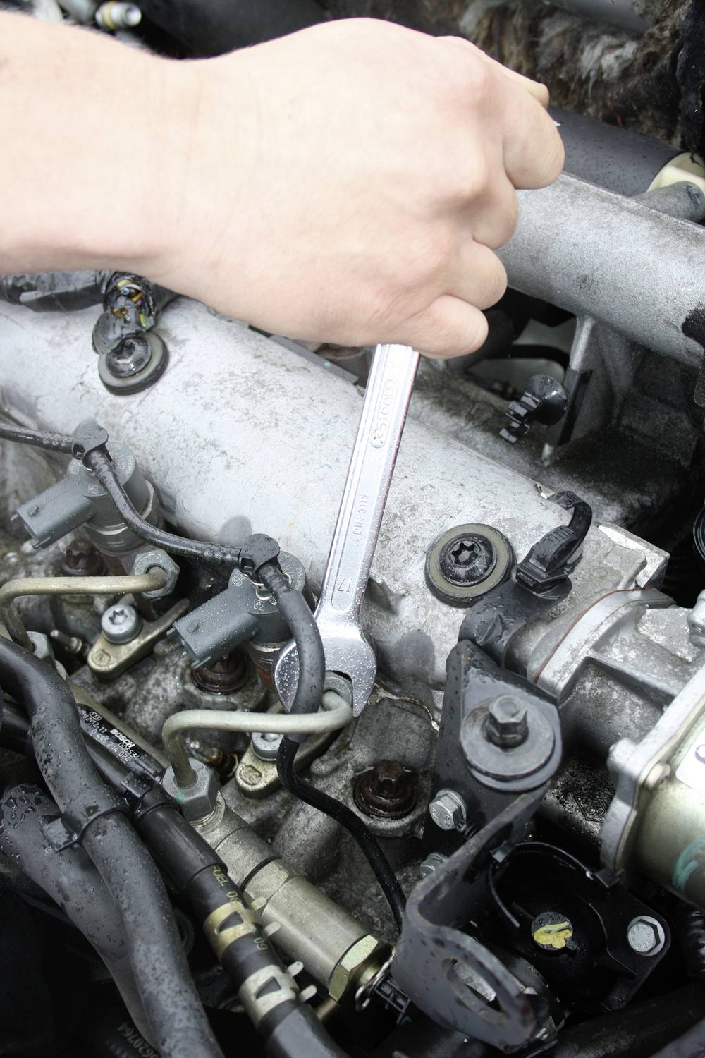 Egy kéz egy villáskulcsot tart, miközben egy motornál dolgozik. A motor több csövet és kábelt mutat.