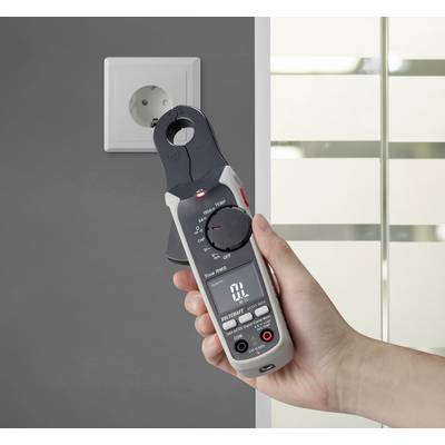 Egy kéz egy digitális multiméterrel egy szürke fal melletti konnektor közelében áll. A készülék kijelzőjén '0 L' látható.