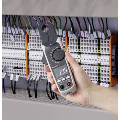 Egy kéz digitális feszültségmérőt tart egy kapcsolószekrény előtt, amelyben sok kábel fut. A kijelzőn '239' látható.