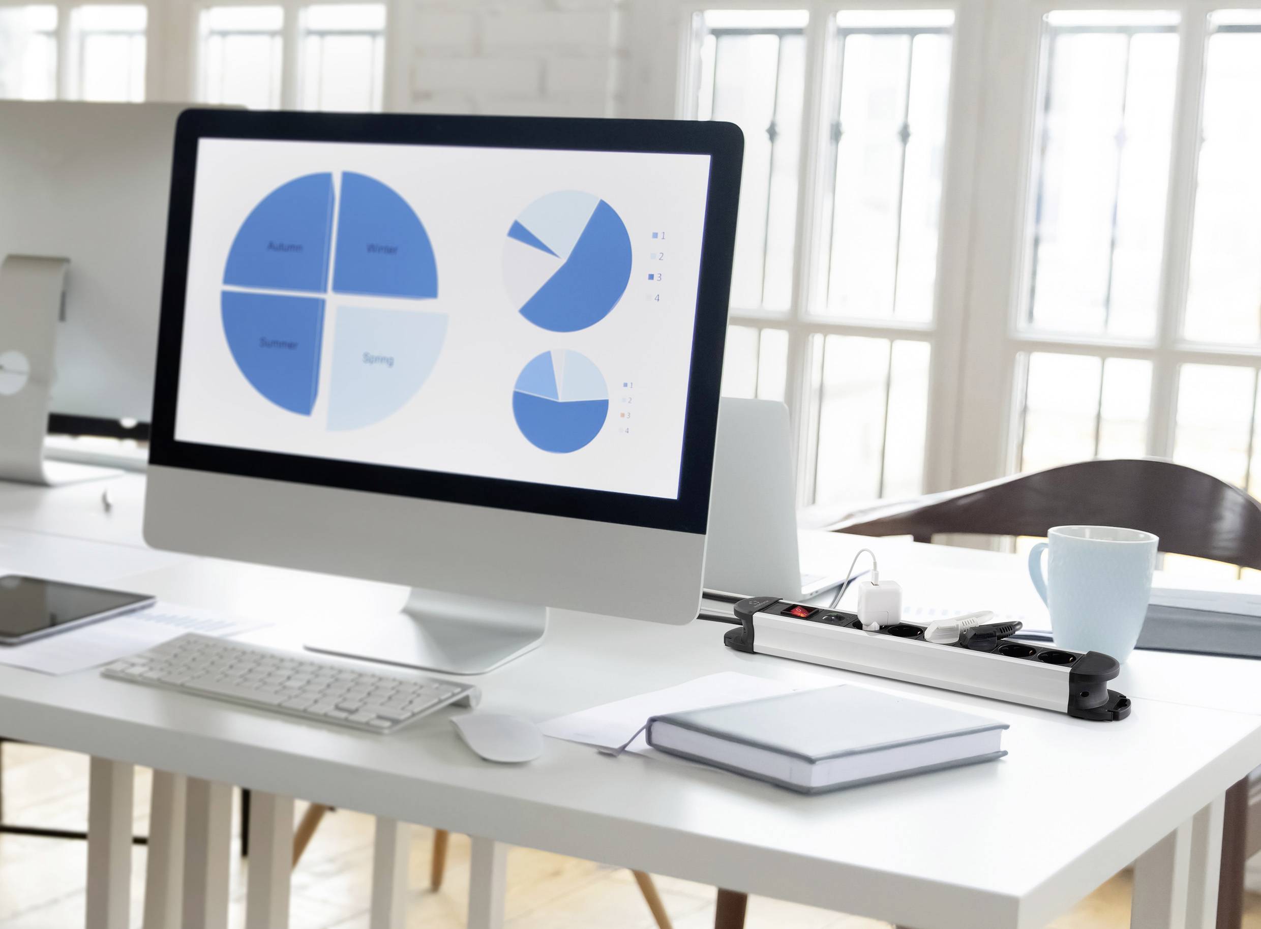 Egy számítógépes képernyő három kék kördiagramot mutat, amelyek adatokat tartalmaznak 'Marketing', 'Értékesítés', 'Pénzügy' és 'Üzemeltetés' témakörökben. Irodai környezet látható a háttérben.