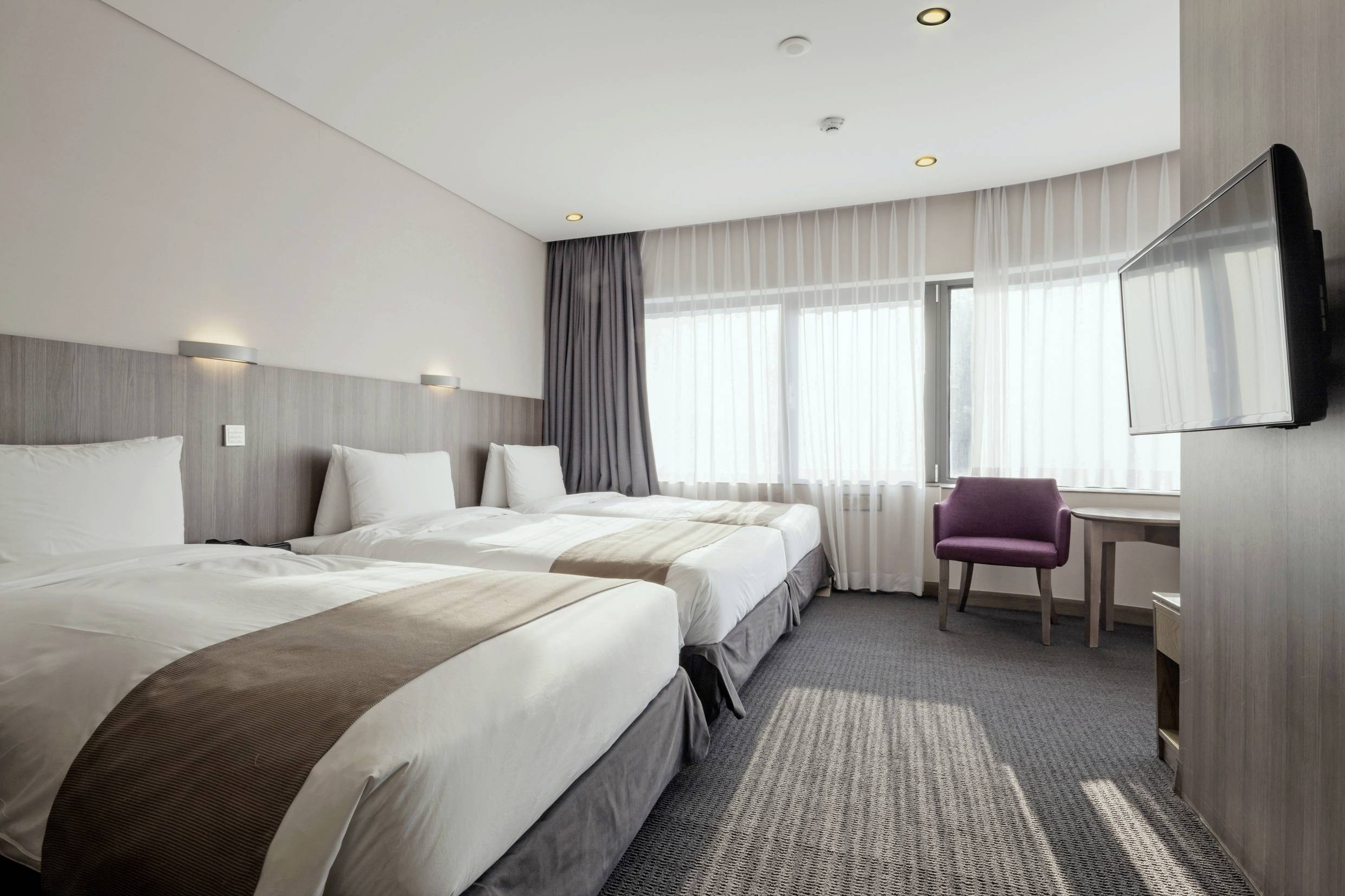 Három egyszemélyes ágy egy modern hotelszobában, íróasztallal és a fal melletti televízióval. Világos függönyök engedik be a nappali fényt.