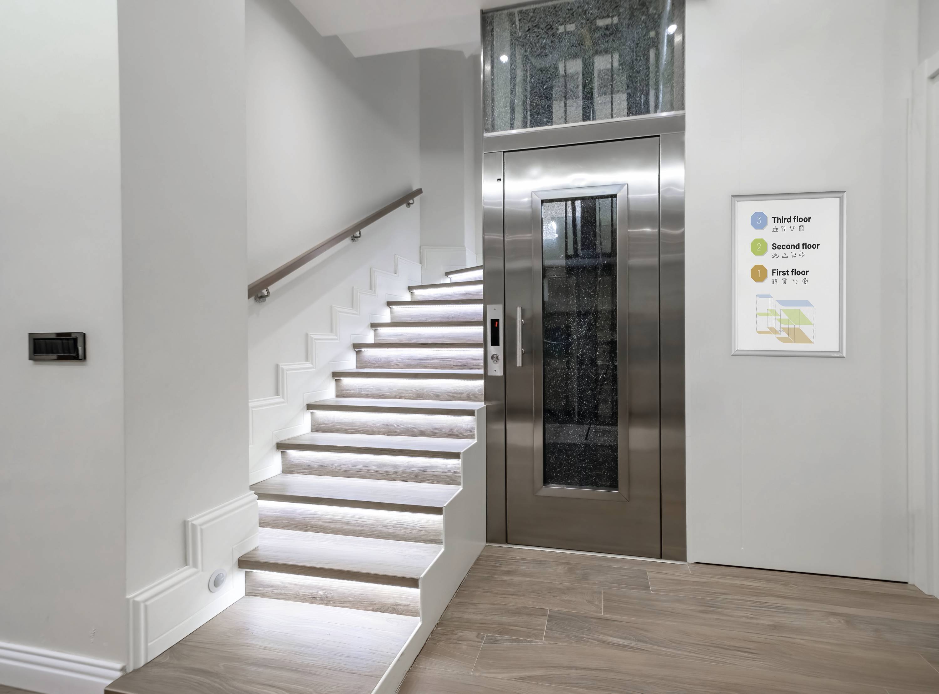 Lépcsőház modern lépcsővel és felvonóval. A falon egy tábla függ, amely színnel jelölt szintinformációkat tartalmaz.