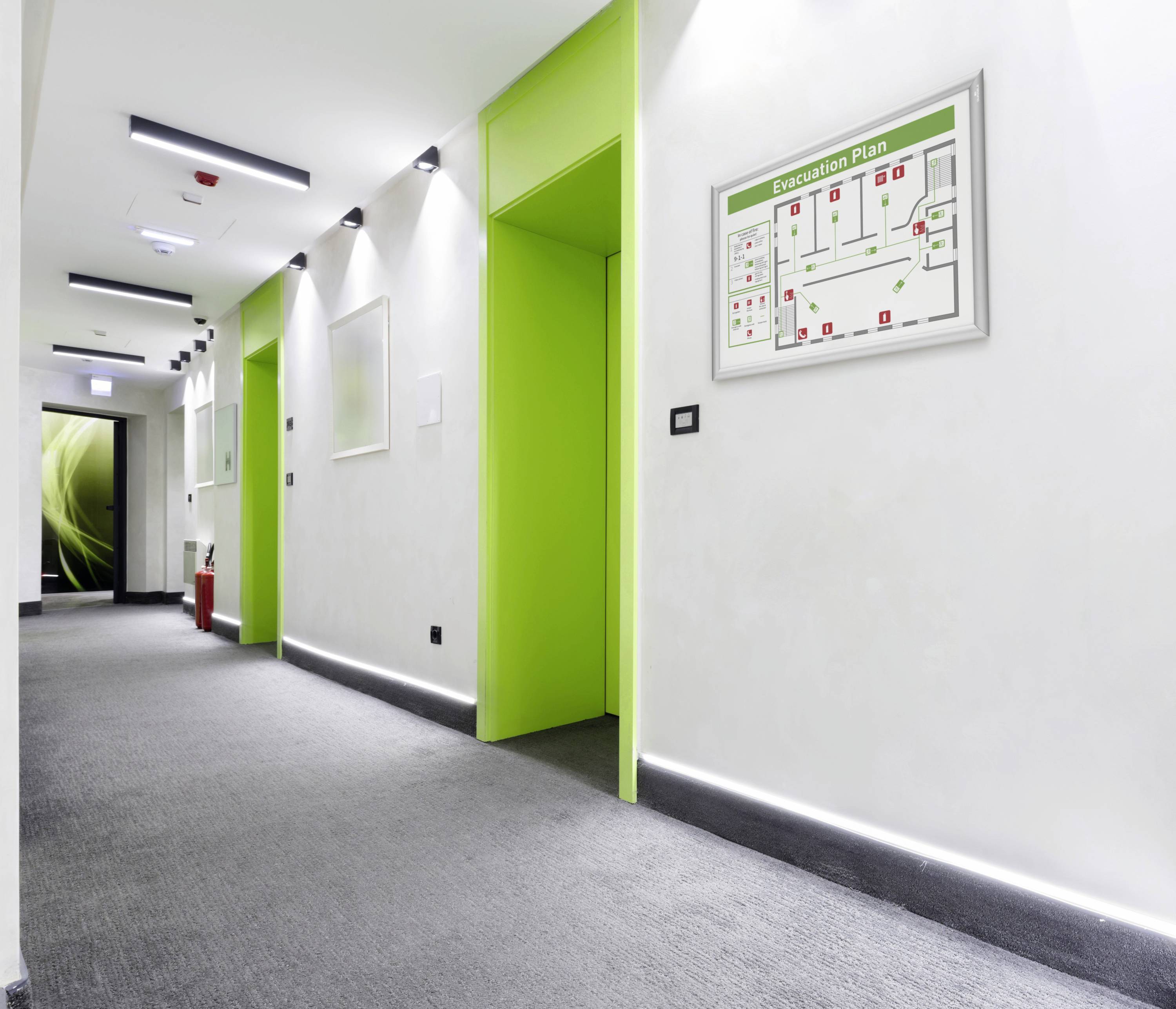 Egy világos folyosó zöldre festett ajtókkal és egy fali kiürítési tervvel. Baloldalt hátul egy tűzoltókészülék áll.