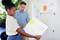 Egy nő egy férfinak mutat egy plakátot diagramokkal és feljegyzésekkel. Egy projektről beszélgetnek. Növények vannak a háttérben.