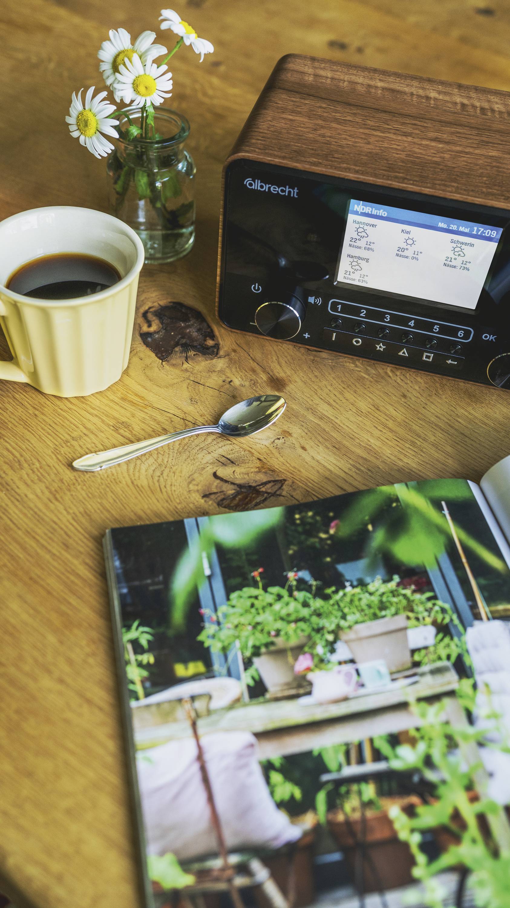 Egy csésze kávé, egy rádió, egy kanál, egy váza virágokkal és egy kinyitott könyv növényfotókkal egy fa asztalon fekszenek.