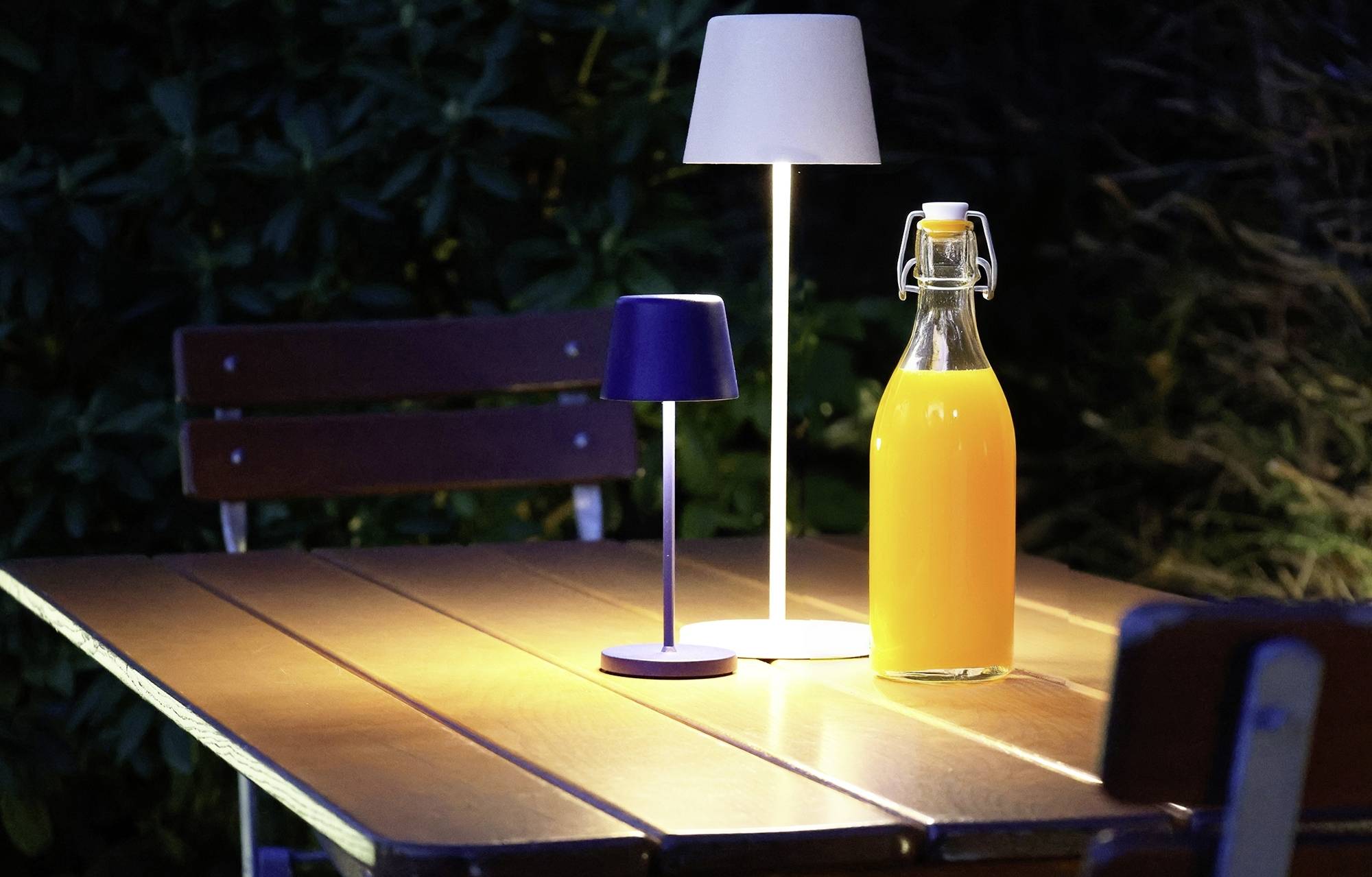 Egy narancssárga folyadékkal teli megvilágított üvegpalack áll egy faasztalon, két kisebb lámpa között; a háttérben bokrok láthatók.