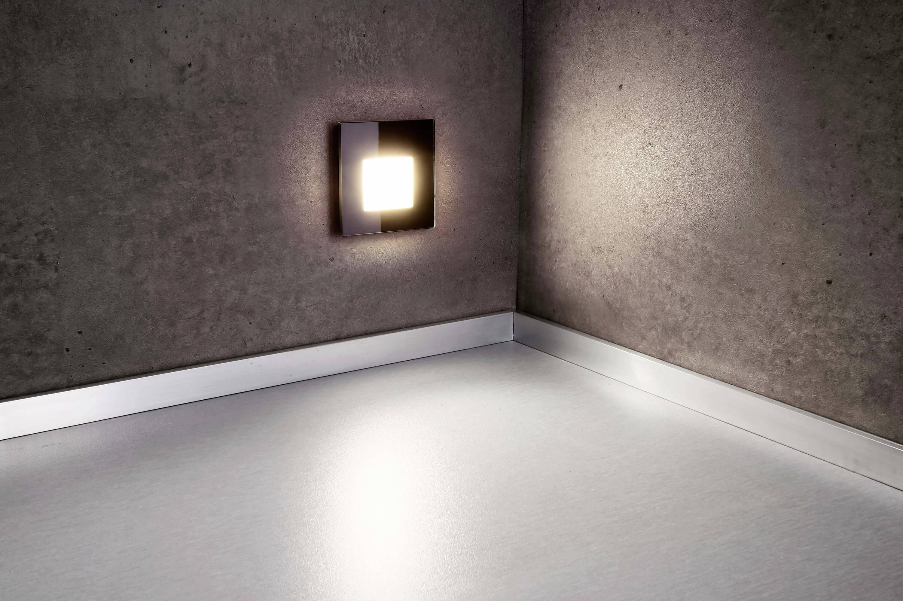 Modern fali lámpa dizájn egy minimalista térben. A téglalap alakú lámpa egy világos padlón egy sarkot világít meg.