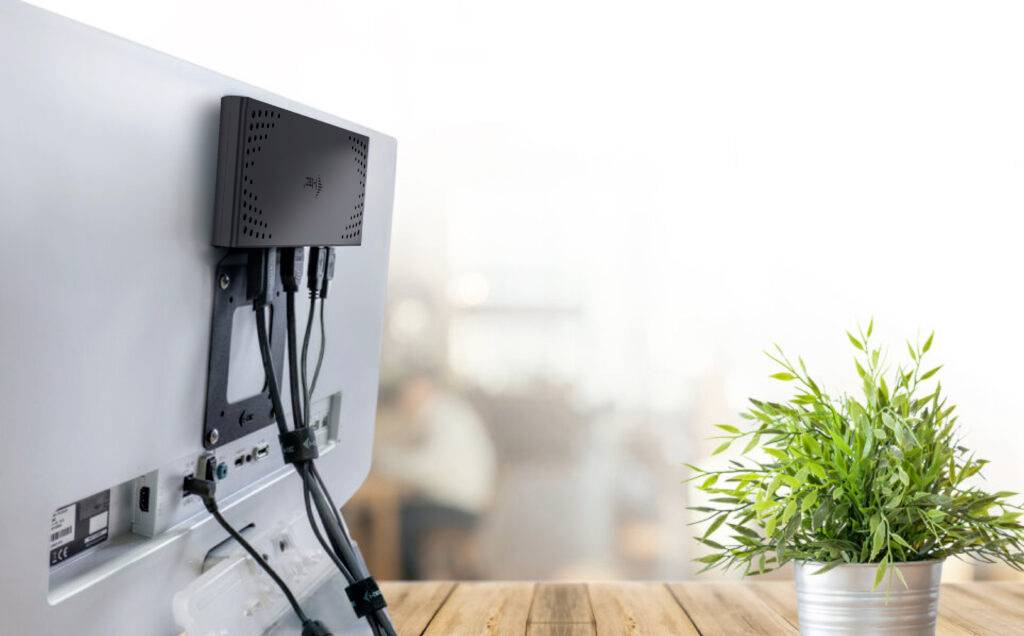 Egy számítógép képernyő hátulról nézve, több csatlakoztatott kábellel, egy faasztalon áll egy zöld cserépnövény mellett.