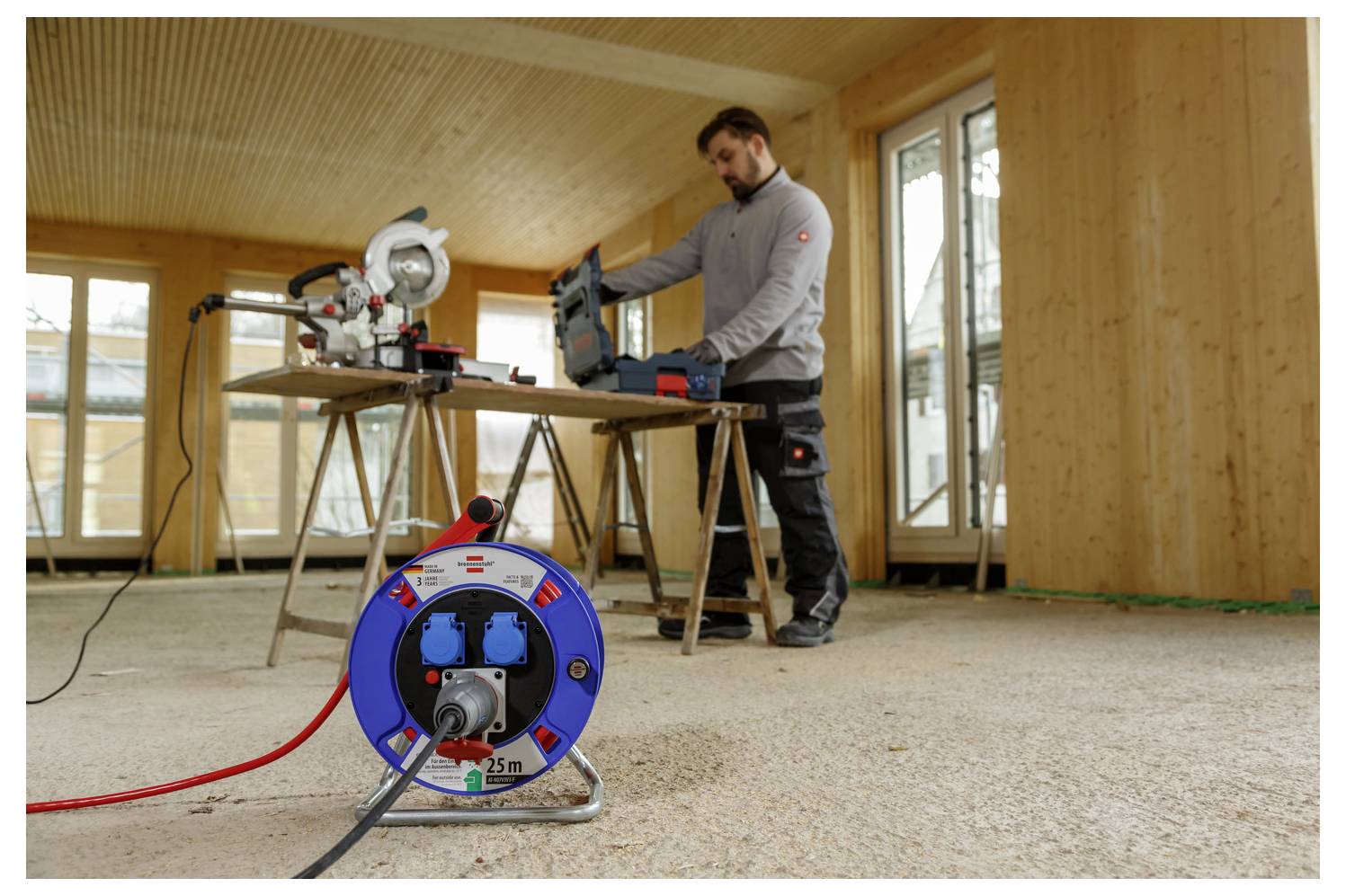 Egy férfi elektromos fűrésszel dolgozik egy munkaasztalnál egy befejezetlen szobában, az előtérben egy kék kábeltekercsel.