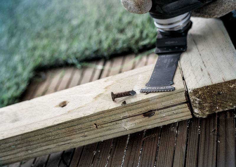 Elektromos kéziszerszám fa deszkát vág egy rozsdás csavar közelében egy fa padlón. A háttérben műfű látható.