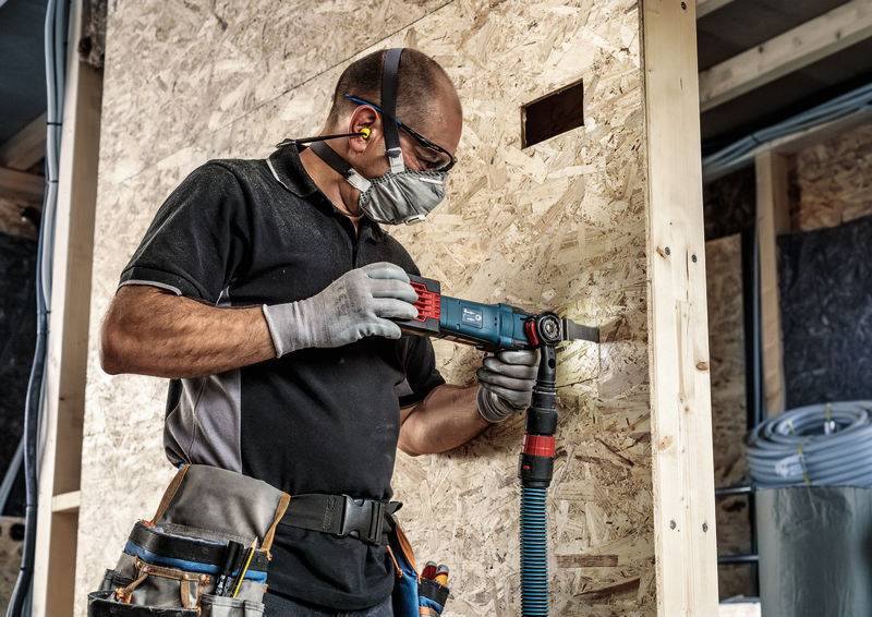 Egy férfi védőszemüvegben, maszkban és fülvédőben fúr egy forgácslemezes falba. Szerszámövet és munkakesztyűt visel.