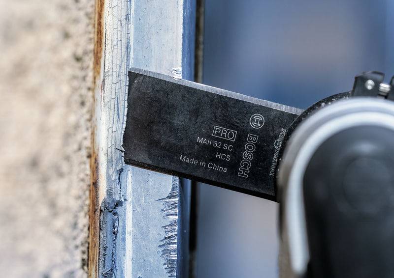 Kézi szerszám vág tömítőanyagot egy ablakkeret szélén. A pengén a 'Bosch' felirat látható. Közeli felvétel.