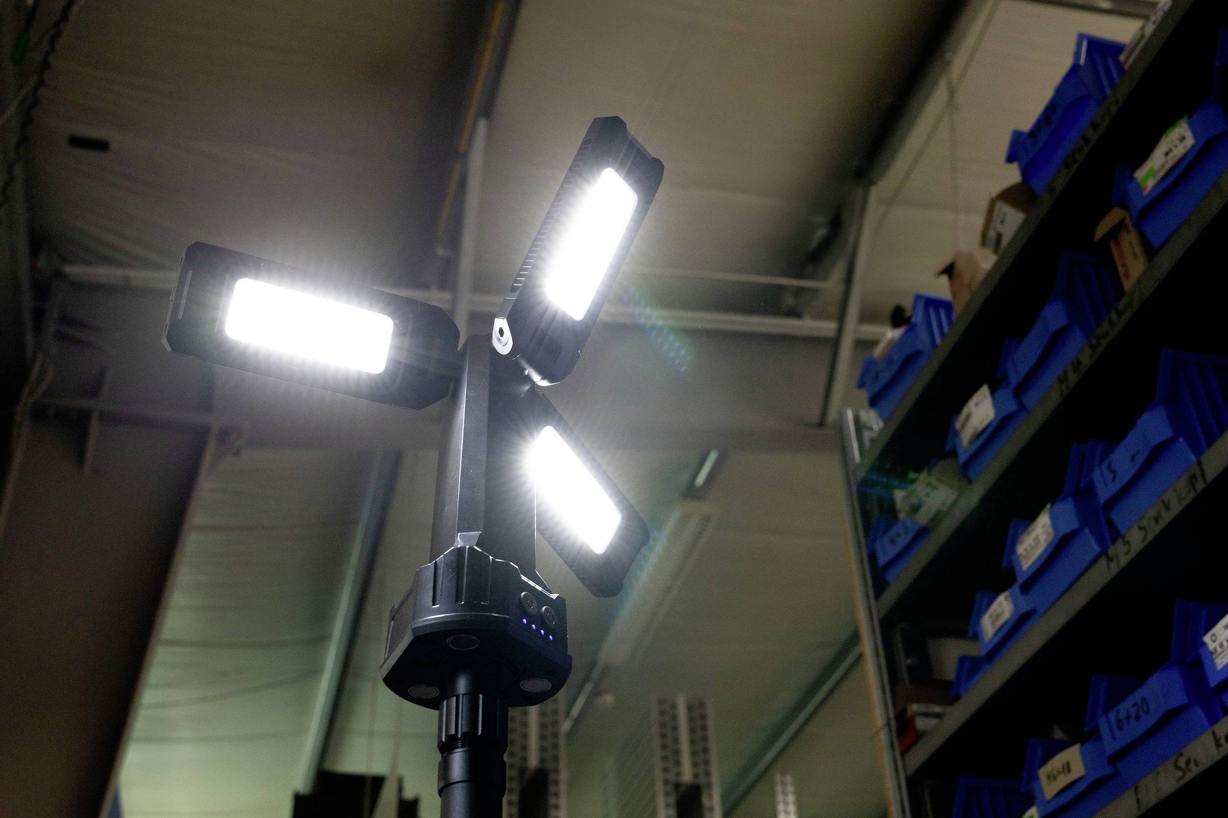 Egy világos, négykaros LED-reflektor világít egy raktárhelyiségben, amelynek hátterében kék polcok vannak.
