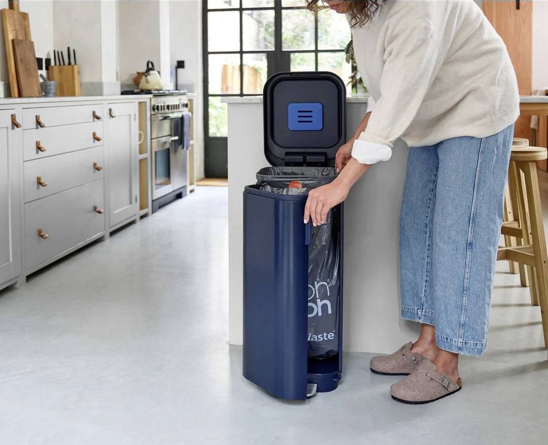 Egy személy egy modern konyhában egy nyitott tetejű sötétkék szemétkosárba dob valamit.