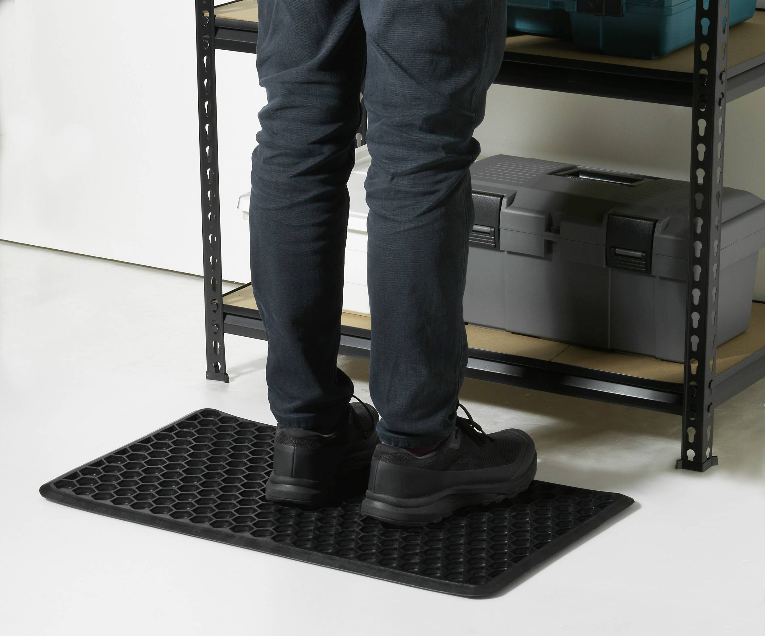 Egy személy áll egy méhsejt mintás, csúszásmentes gumiszőnyegen egy szerszámosdobozokkal teli polc előtt.