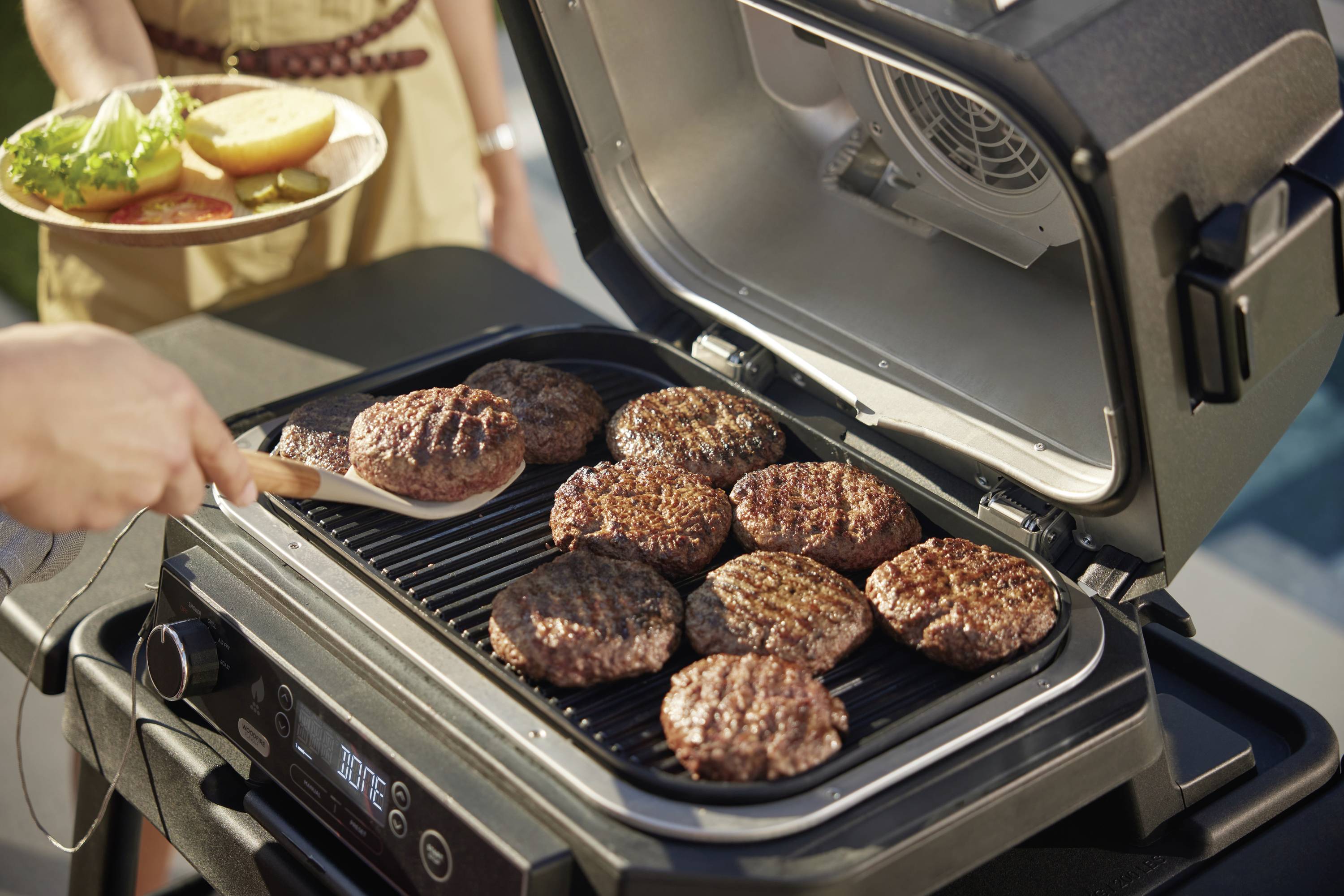 Egy személy nyolc hamburger-pogácsát süt egy elektromos grillsütőn a szabadban. Egy második személy egy tányért tart, rajta zsömlékel és salátával.