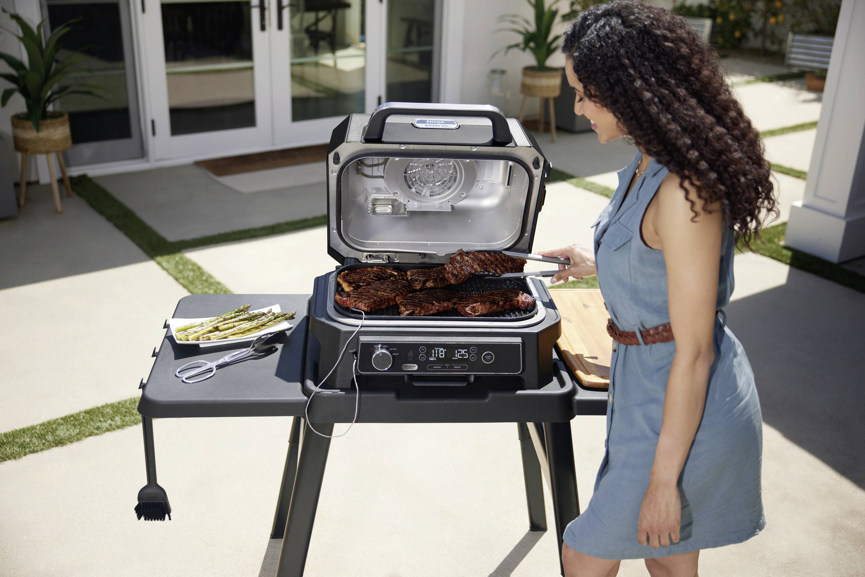 Egy személy húst süt egy elektromos grillsütőn a szabadban. A grill jobb oldalán grillezett spárga fekszik egy tányéron.